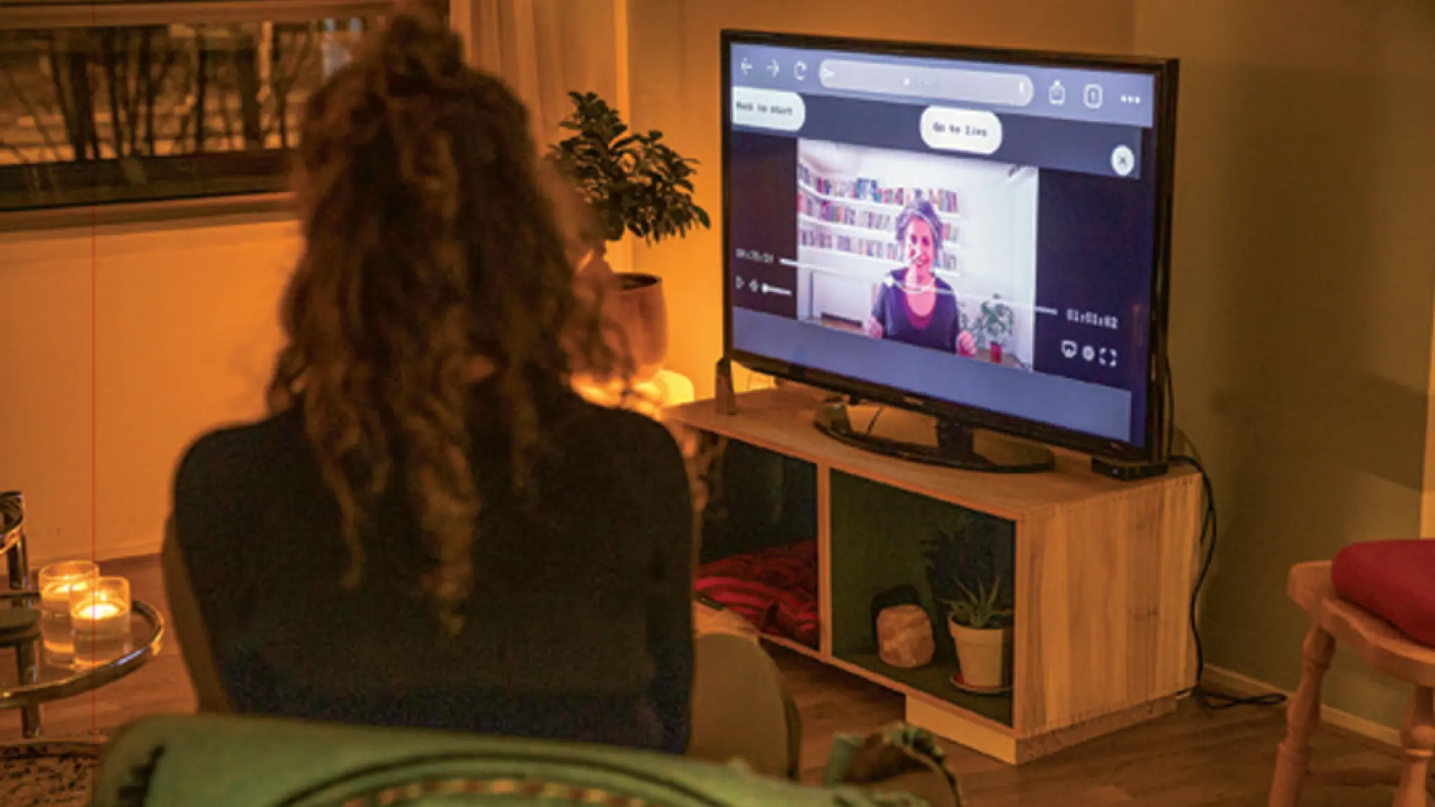 Een vrouw zit in een gezellige woonkamer en kijkt naar een televisie waarop een videogesprek te zien is met een andere vrouw. De kamer is sfeervol verlicht met kaarsen en planten.