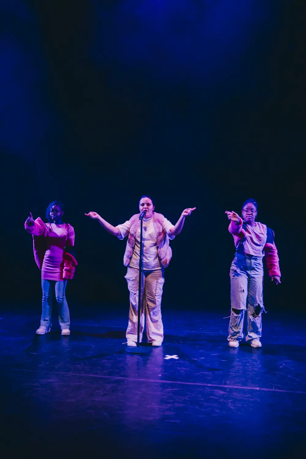 Foto van meerdere deelnemers aan de Jeugdtheaterschool Zuidoost. Drie meisjes staan naast elkaar op het podium. De middelste praat in een microfoon en wijst daarbij de anderen aan. De meisjes links en rechts aan de buitenkant wijzen met hun rechterhand naar voren.