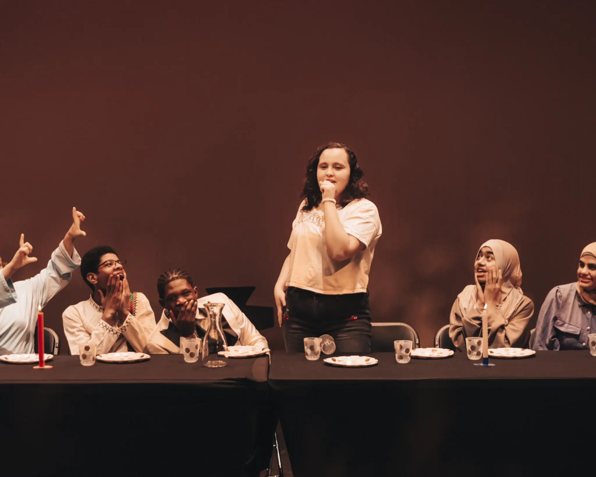 Foto van meerdere deelnemers aan de Jeugdtheaterschool Zuidoost. Ze zitten aan een lange tafel naast elkaar. Een meisje in het midden staat rechtop en doet alsof ze zingt of praat in een microfoon. De anderen kijken vol bewondering naar haar.