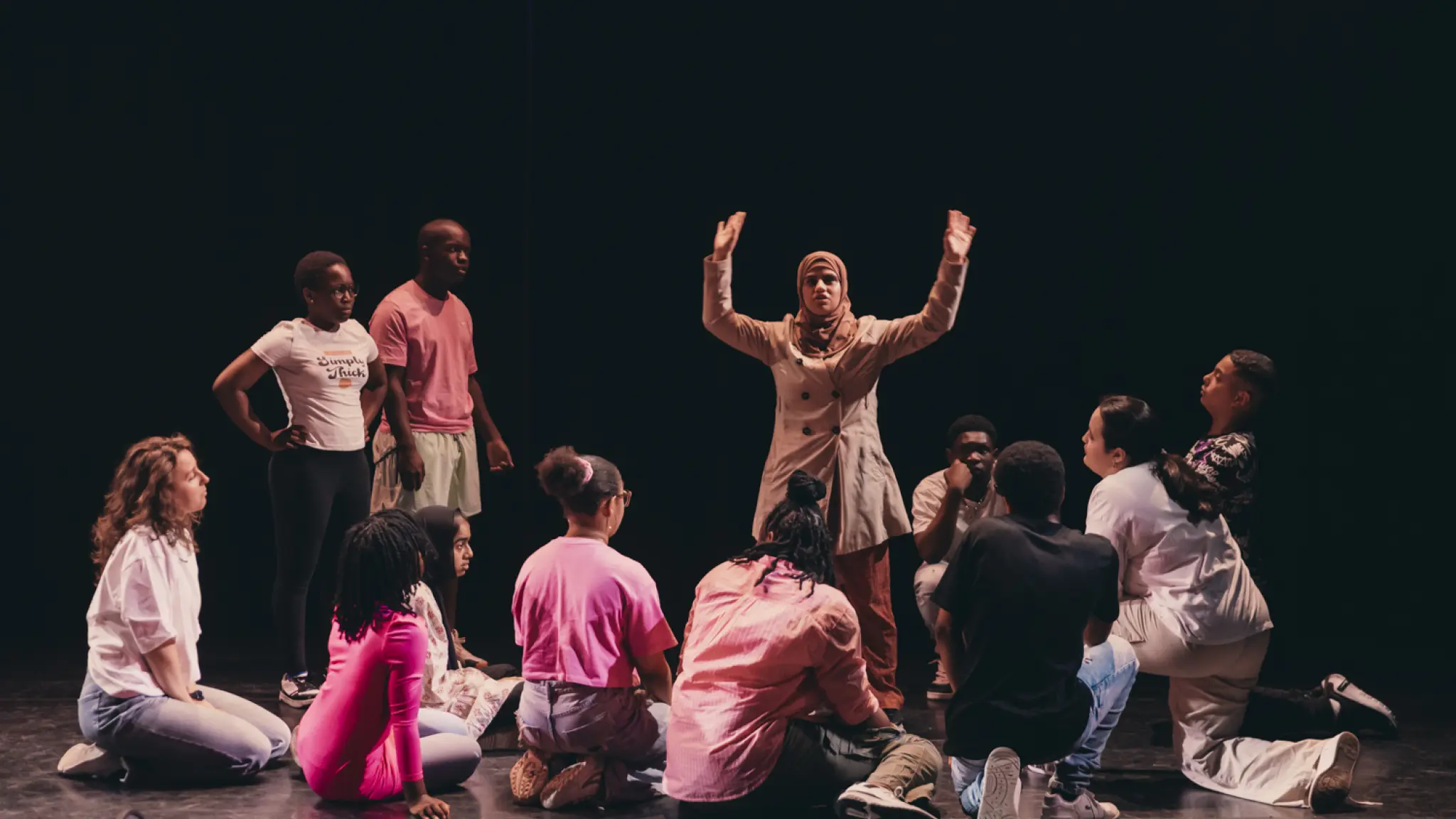 Foto van meerdere deelnemers aan de Jeugdtheaterschool Zuidoost. Een meisje met hoofddoek staat in het midden van het podium met haar handen in de lucht. De andere deelnemers zitten, staan en knielen op het toneel om haar heen, met het gezicht naar haar gericht.