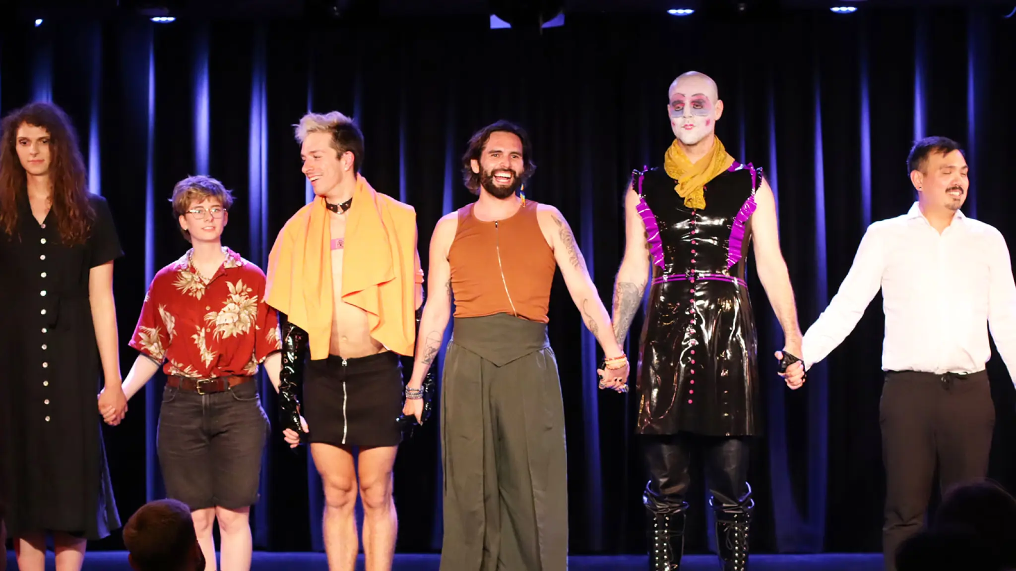 Luis Bracamontes staat in het midden van enkele performers op het podium. Ze staan op een rij en houden elkaars handen vast. Het publiek geeft applaus.