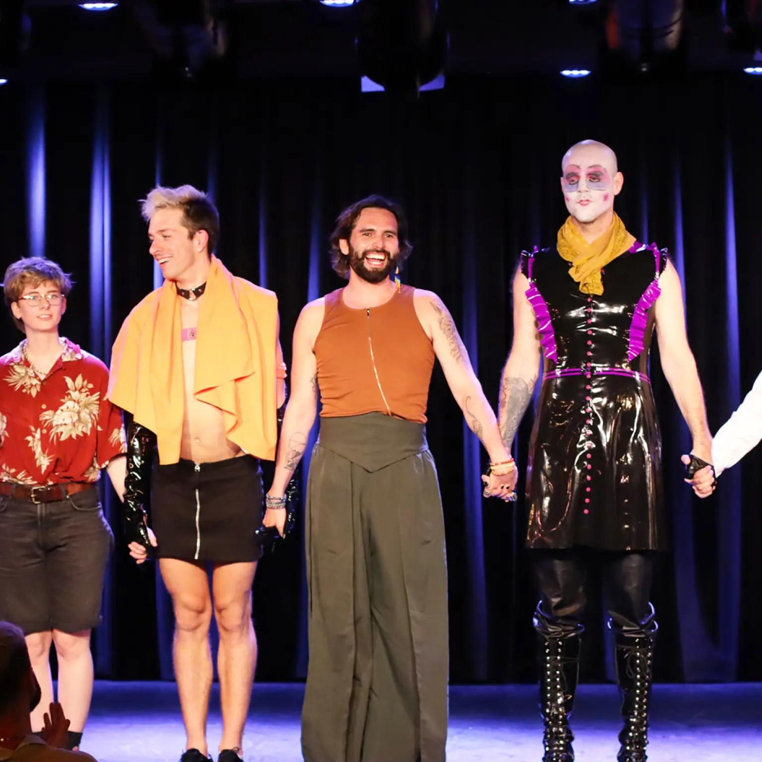 Luis Bracamontes staat in het midden van enkele performers op het podium. Ze staan op een rij en houden elkaars handen vast. Het publiek geeft applaus.