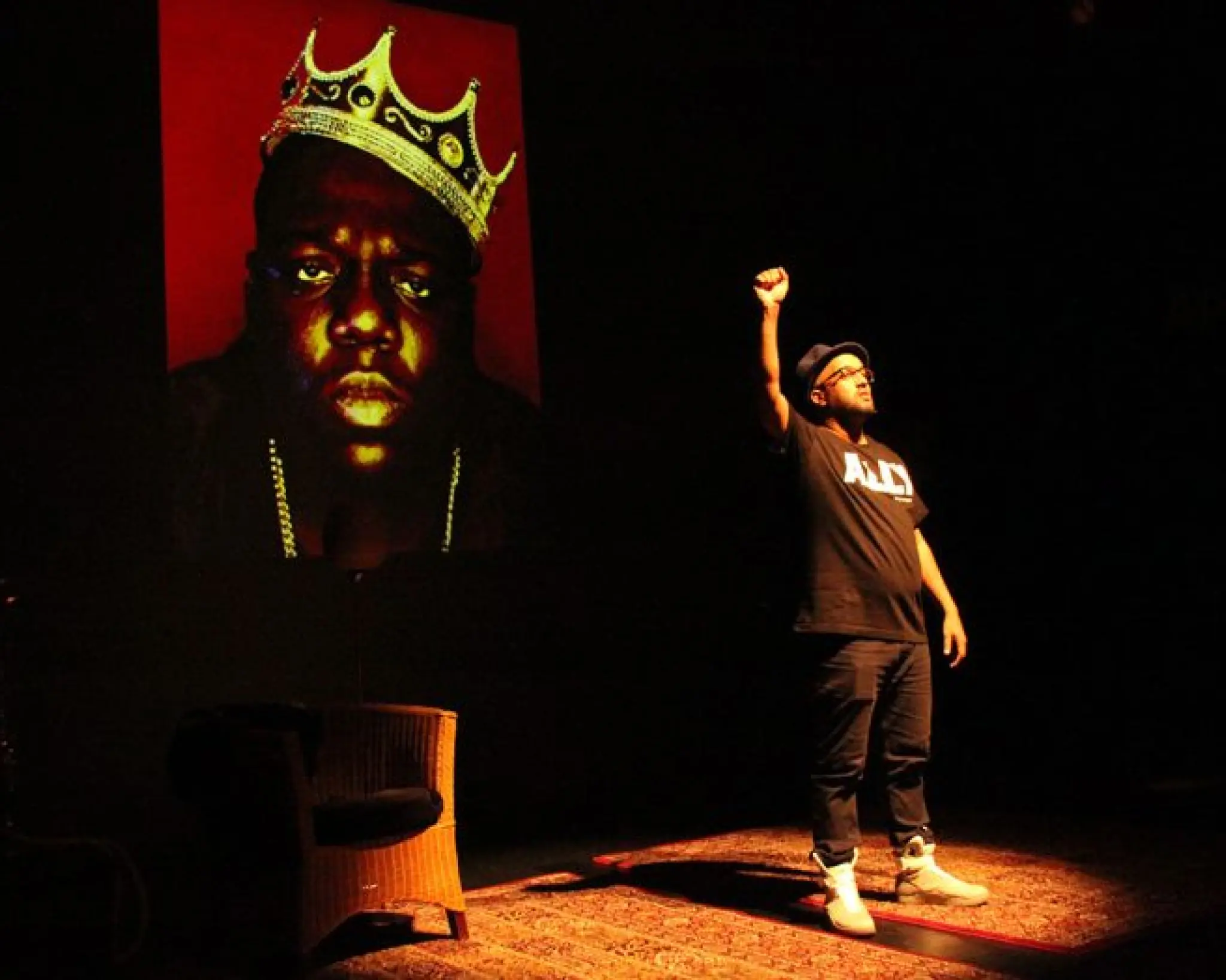 Mo Hersi steekt zijn rechterarm met gebalde vuist de lucht in. Op de achtergrond is een foto te zien van rapper The Notorious B.I.G. (Biggie Smalls).