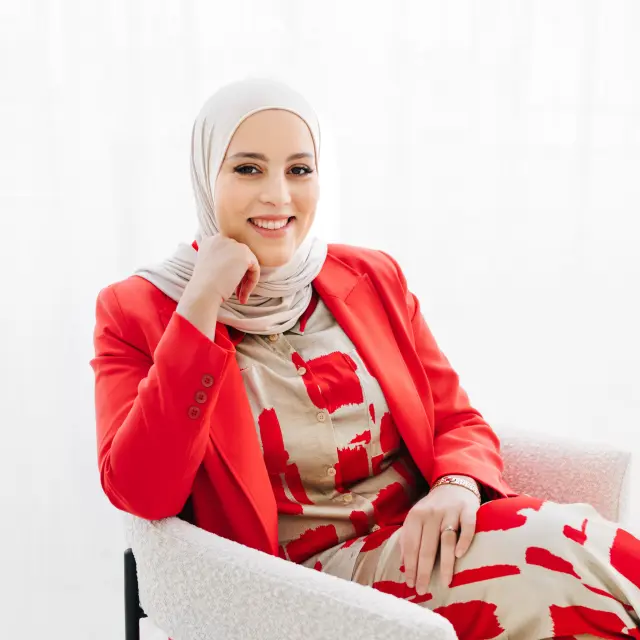 Hanan draagt een beige met rood pak en een rood jasje eroverheen Ze draagt een beige hoofddoek en lacht vriendelijk naar de camera.