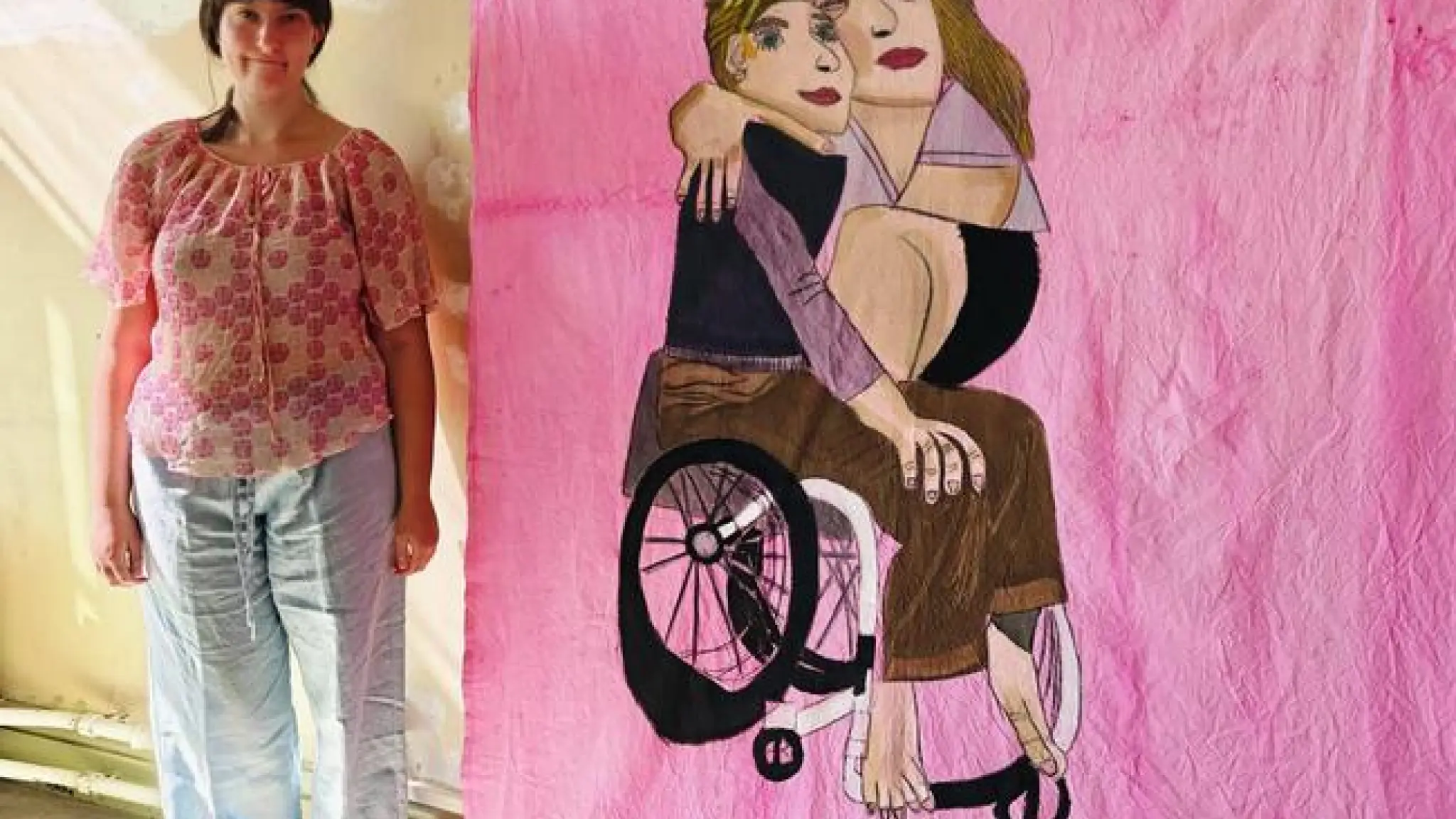 Een vrouw staat naast een groot kunstwerk op roze stof waarop twee mensen zijn afgebeeld, waarvan één in een rolstoel zit terwijl de ander op schoot zit en hen omhelst.