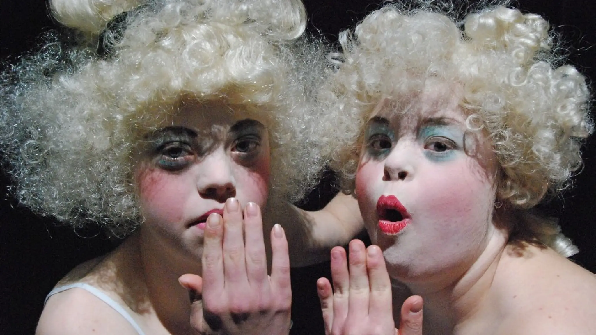 Twee acteurs van KamaK zijn uitgedost met witte pruiken en make-up. Ze houden een hand voor hun mond alsof ze verbaasd zijn.