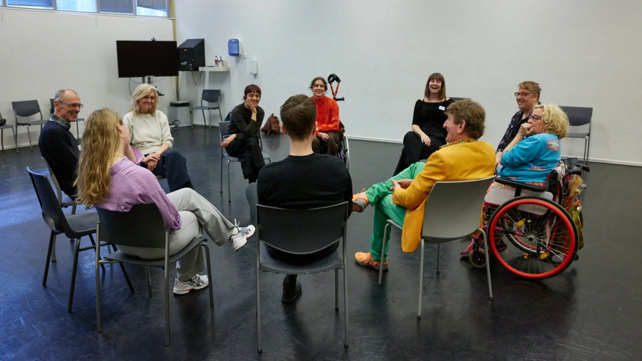 Een diverse groep mensen, waaronder een persoon in een rolstoel, zit in een kring in een lichte ruimte en voert een gesprek of groepsactiviteit; iedereen lijkt betrokken en ontspannen.