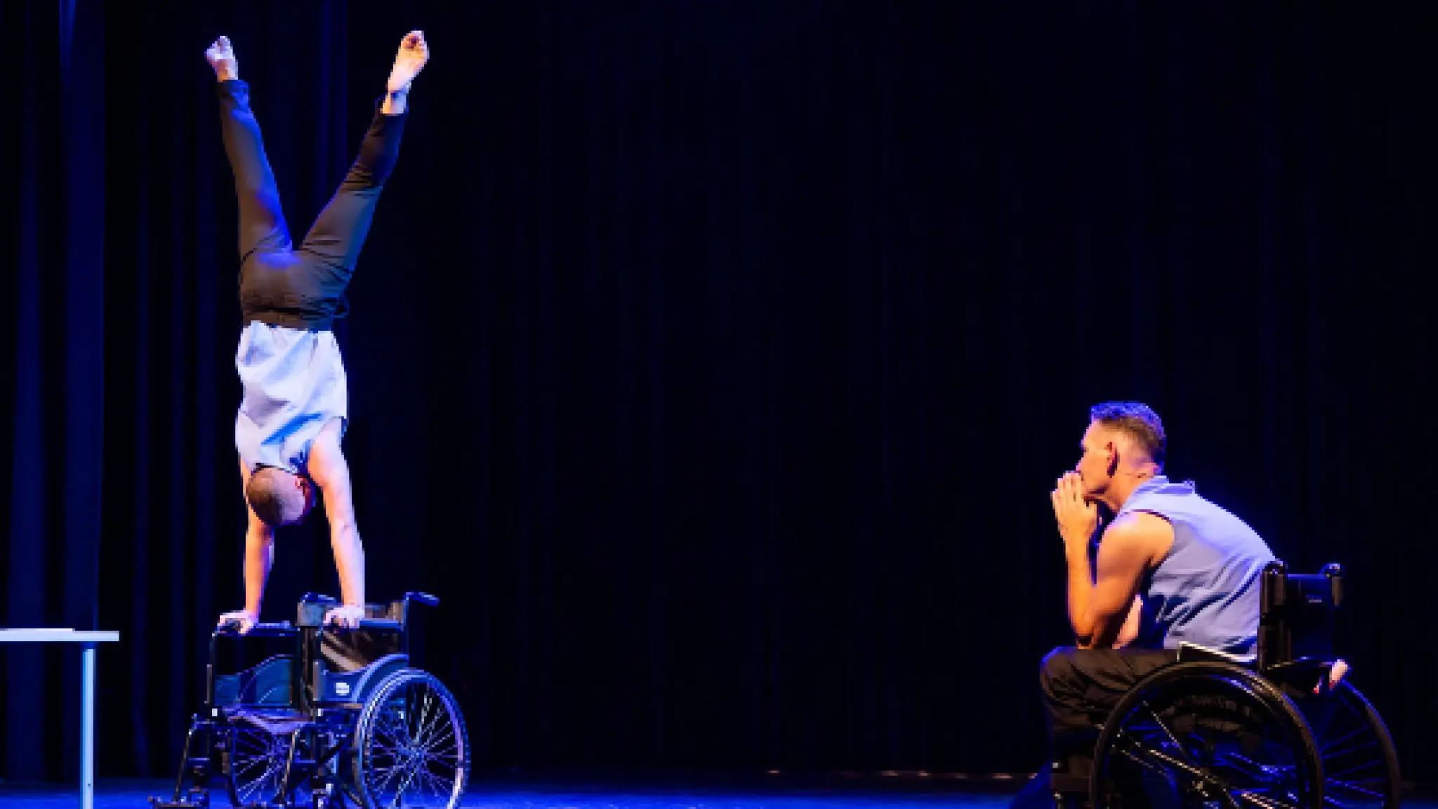 Op een theaterpodium doet een persoon een handstand op de armleuningen van een rolstoel. Rechts kijkt een tweede persoon in een rolstoel aandachtig toe, met de handen voor het gezicht. Beide dragen lichte bovenkleding. De scène is verlicht met blauw podiumlicht tegen een donkere achtergrond, wat de focus legt op de acrobatische uitvoering en de interactie tussen de twee performers.