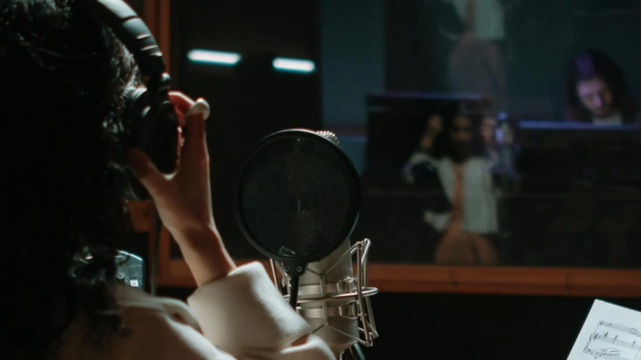 Een persoon met een hoofdtelefoon zit in een opnamestudio achter een microfoon met popfilter. Door het raam van de opnamecabine zijn vaag andere personen zichtbaar. Op de voorgrond ligt een blad met bladmuziek. De ruimte is sfeervol verlicht en straalt een professionele studiosetting uit.