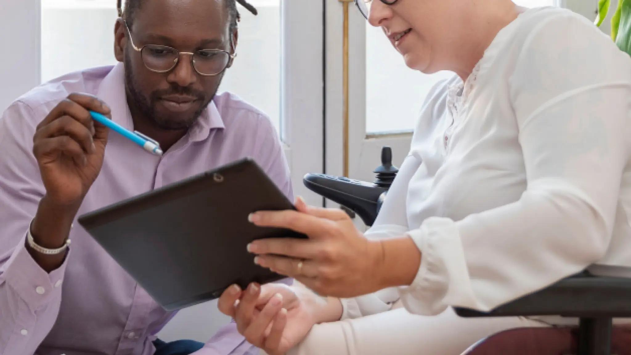 Twee personen zitten naast elkaar in gesprek en bekijken samen informatie op een tablet. De persoon links wijst met een pen naar het scherm, terwijl de persoon rechts de tablet vasthoudt vanuit een rolstoel. Op de achtergrond valt daglicht door een raam.