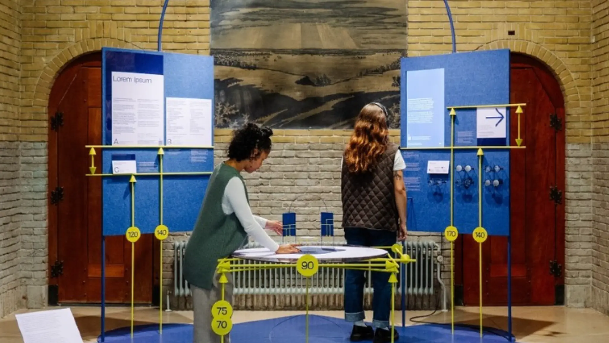 Twee mensen bekijken een interactieve tentoonstelling met blauwe panelen, gele meetlatten en informatieborden in een ruimte met bakstenen muren en een zwart-witfoto aan de muur.
