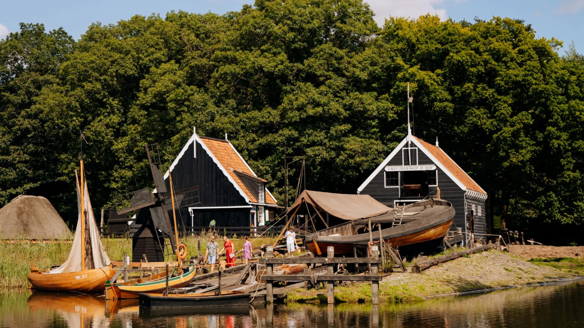 Een historische scheepswerf met houten boten en zwarte huisjes met oranje daken aan het water, omringd door groene bomen, met enkele mensen die het terrein bezichtigen.
