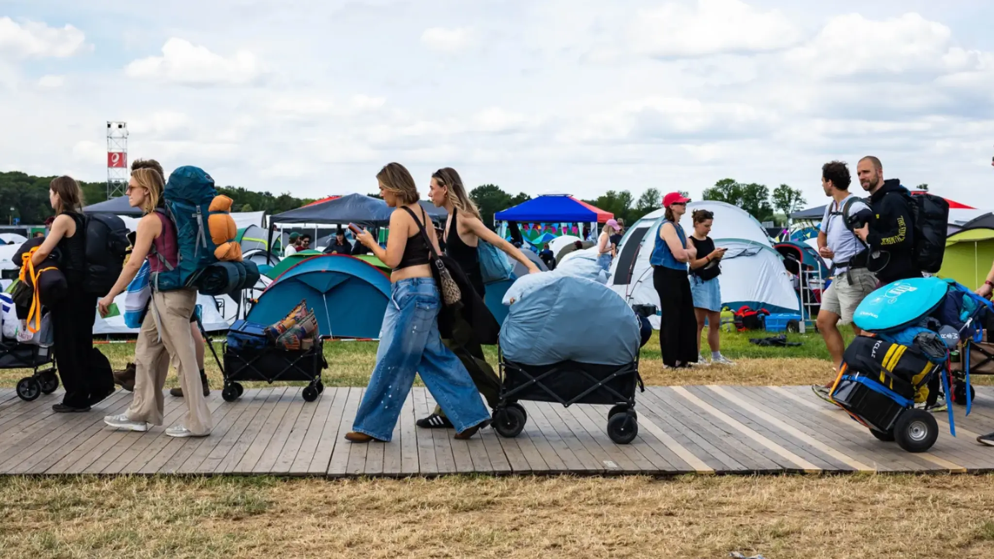Een groep mensen loopt met tassen, rugzakken en wagens vol kampeerspullen over een houten pad op een festivalcamping, terwijl op de achtergrond veel tenten te zien zijn.