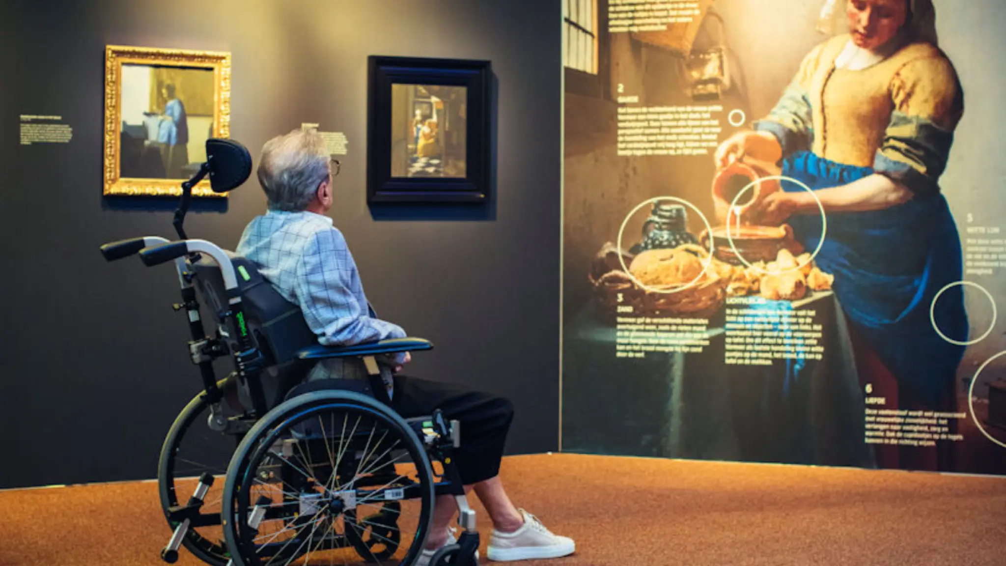Een oudere persoon in een rolstoel bekijkt schilderijen en een grote uitlegwand in een museum, met daarop een afbeelding van "Het Melkmeisje" van Vermeer.