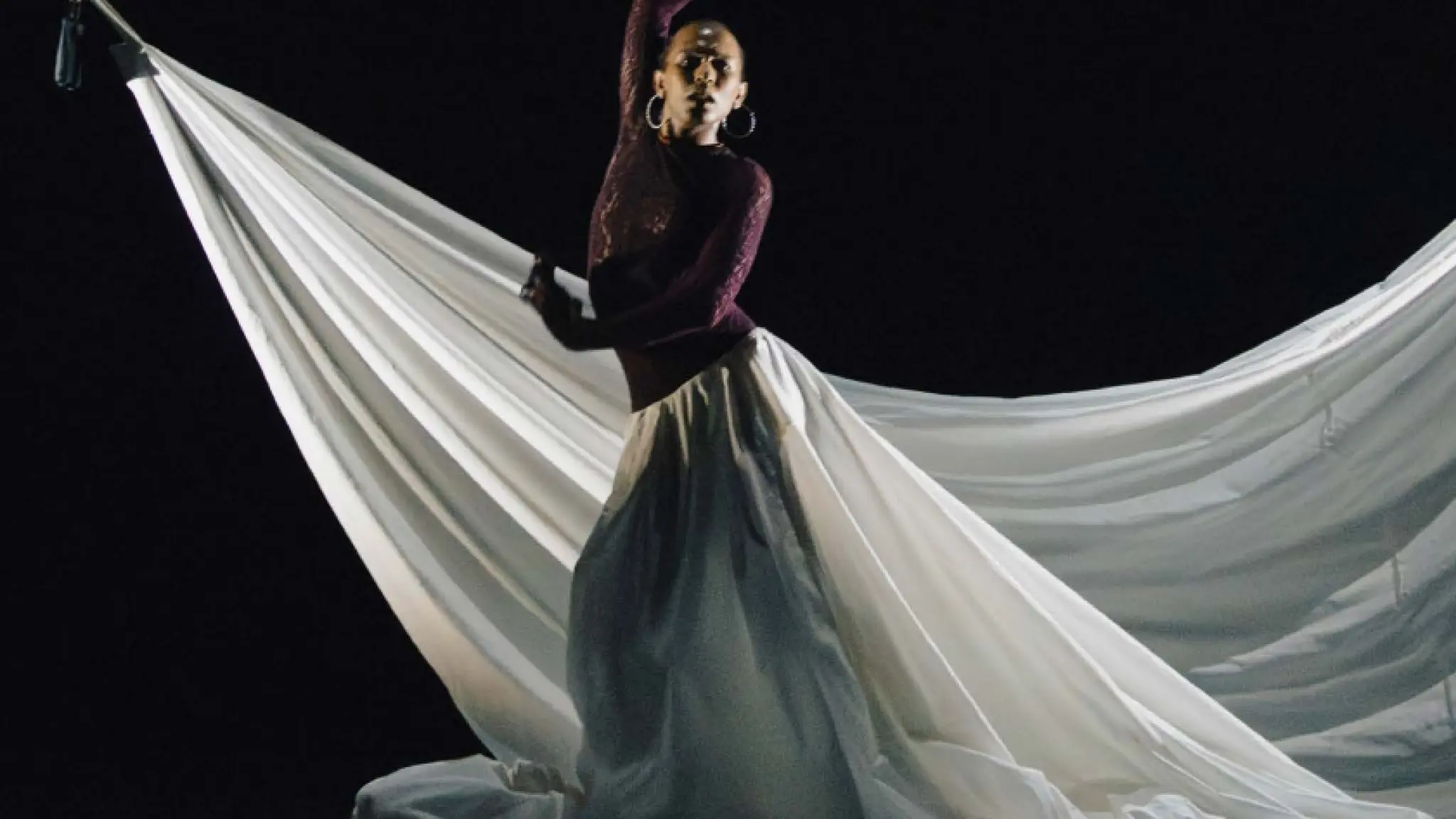 Een danser staat op een donker podium voor een groot, wit doek. Met één arm omhoog en een expressieve houding lijkt de danser midden in een choreografie te bewegen. Het contrast tussen het donkere decor en het lichtgekleurde doek en de wijde rokachtige stof creëert een dramatisch en elegant beeld.