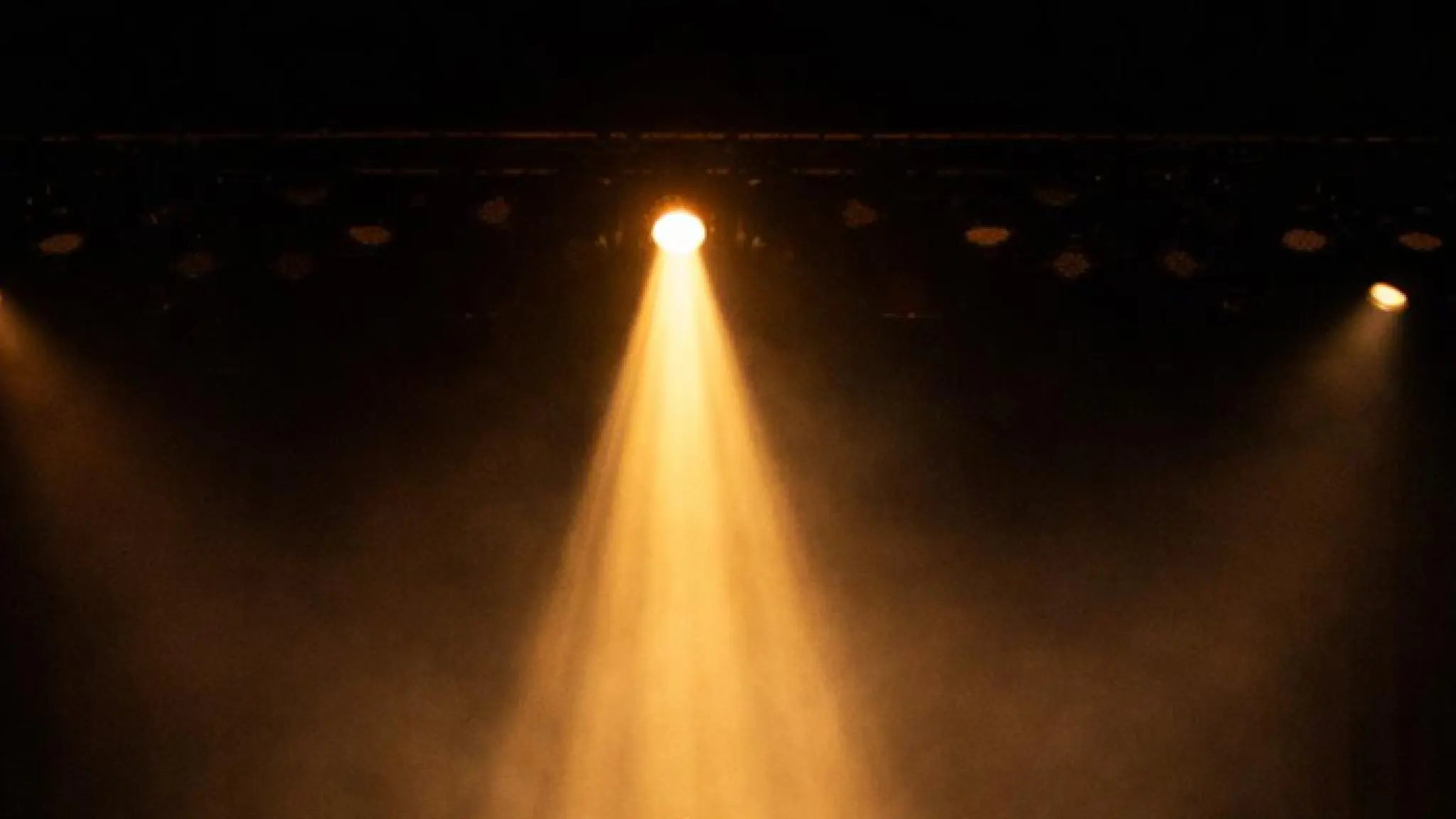 Een donkere theaterscène met een felle gele spot die schijnt op het midden van een leeg podium, omgeven door lichte mist.