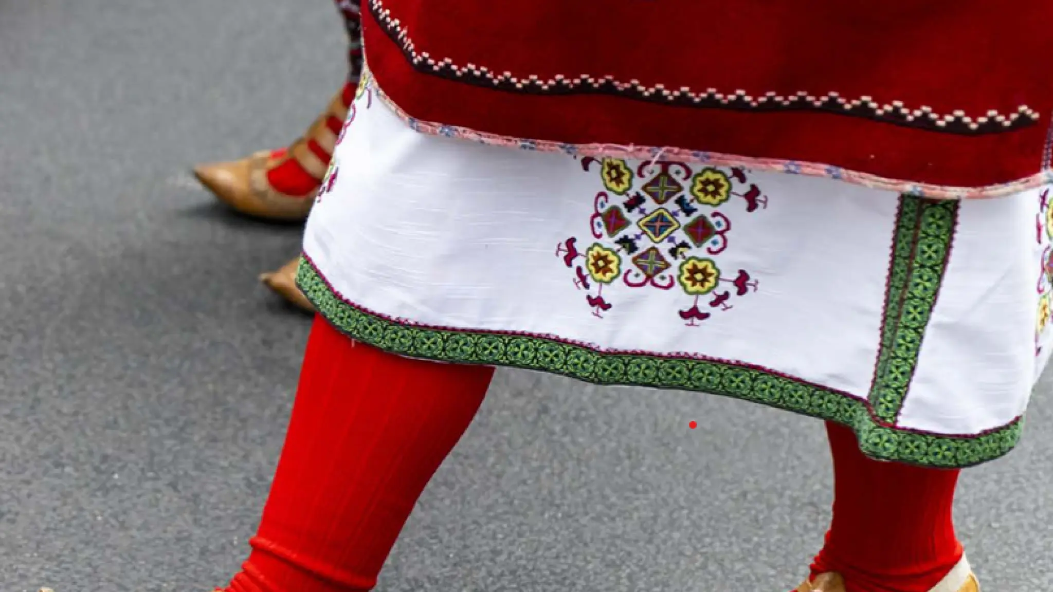 Twee mensen dragen traditionele kleding met felrode sokken, een witte rok met kleurrijke borduursels en leren sandalen met bandjes, terwijl ze op straat lopen.