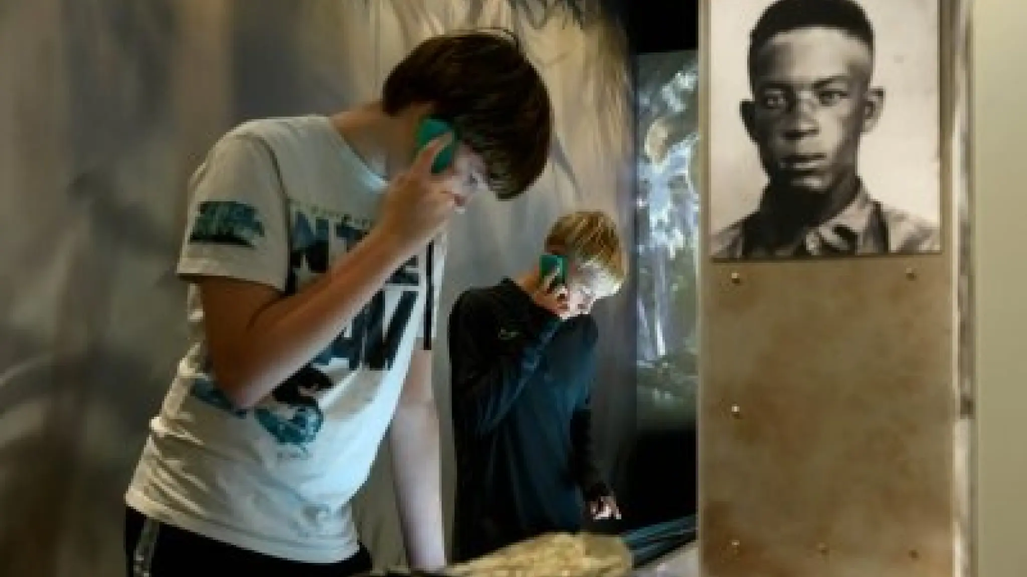 Twee bezoekers staan in een museumruimte en luisteren naar een audiotour via een apparaat dat zij tegen hun oor houden. Op de voorgrond bekijken zij een vitrinekast met objecten, terwijl aan de rechterzijde een zwart-witportret van een man te zien is. De ruimte is gedimd verlicht, waardoor de aandacht uitgaat naar de tentoonstelling en de luisterervaring van de bezoekers.