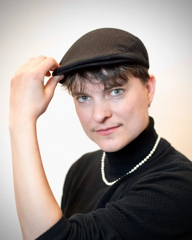 Kir draagt een zwarte coltrui, een parelketting en een baret. Hij kijkt vriendelijk naar de camera terwijl hij wet zijn hand de voorste rand van de baret vastheeft.