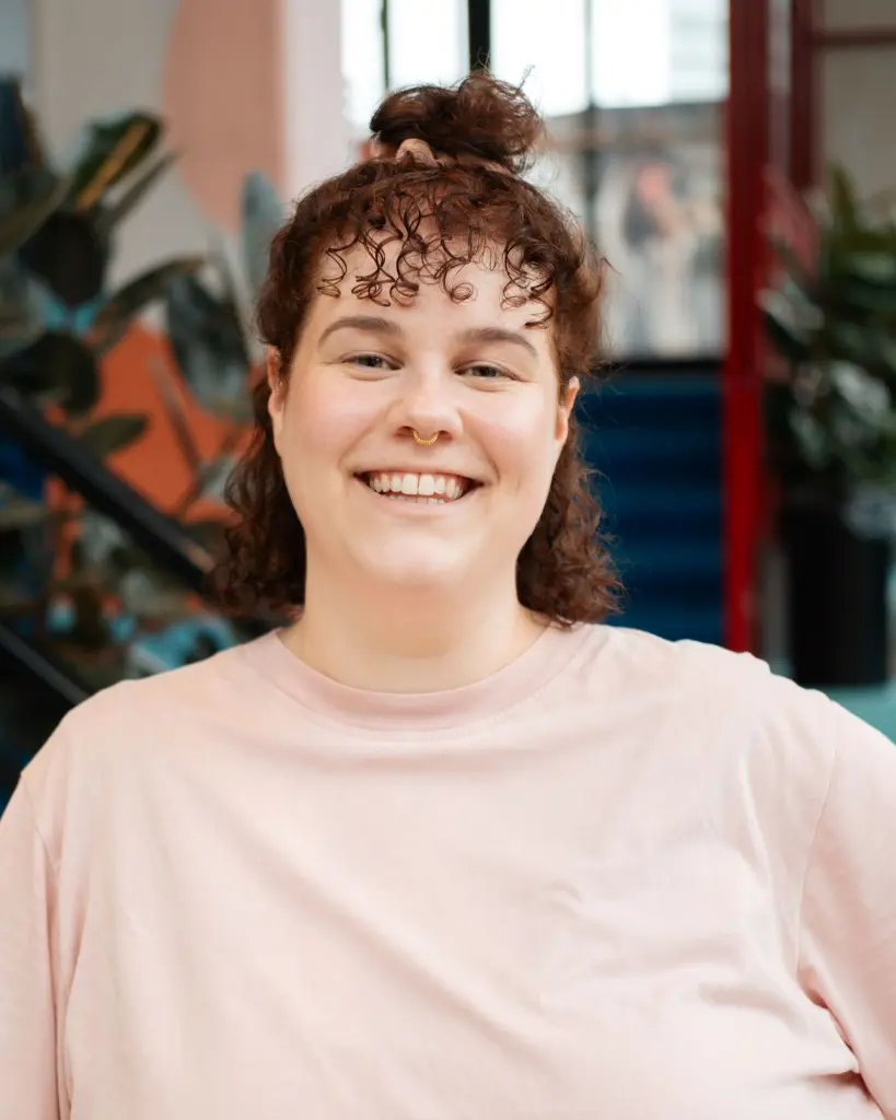 Lisa draagt haar haar in een knot en kijkt lachend naar de camera. Se draagt een lichtroze t-shirt.