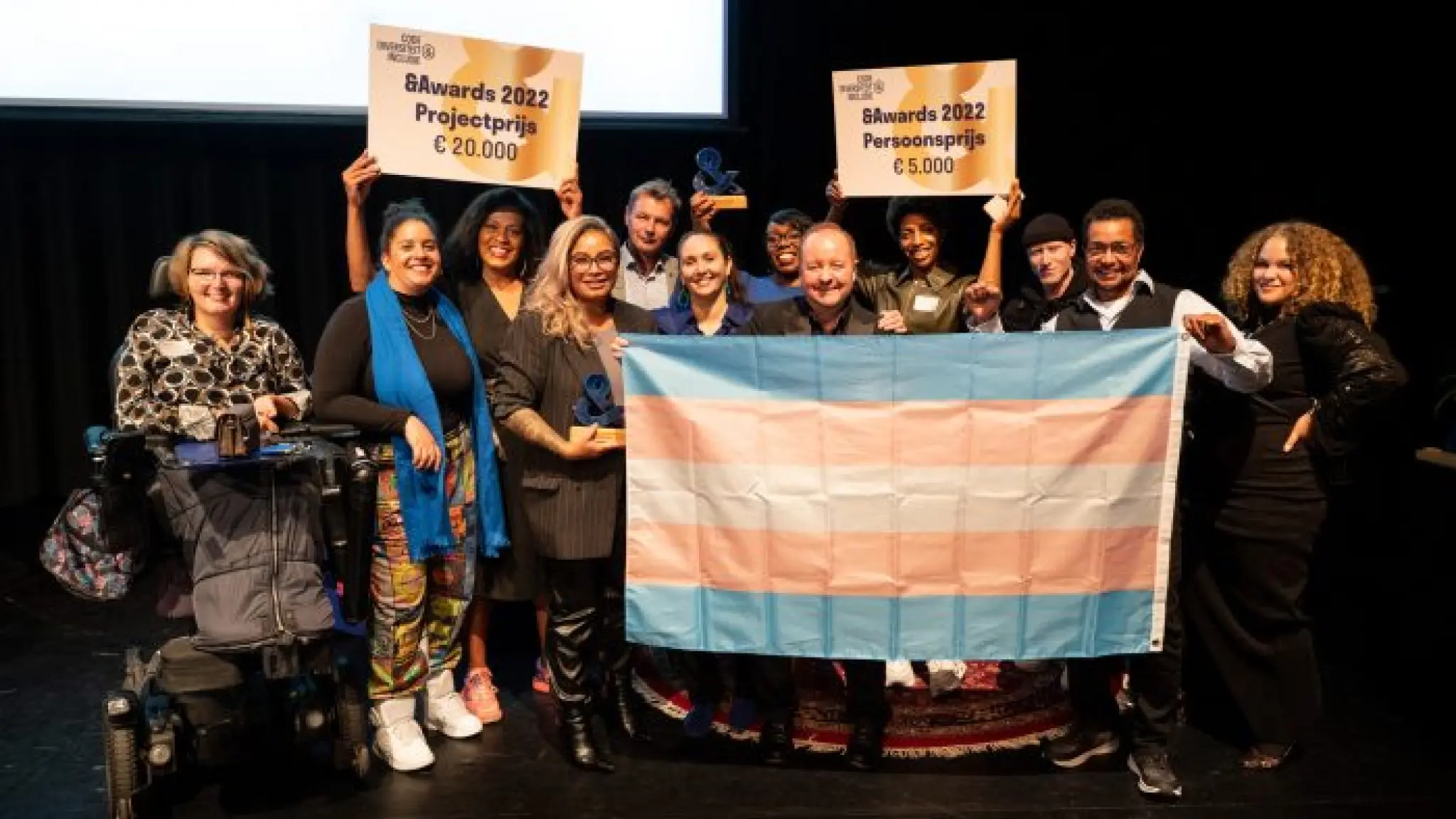 Finalisten en juryleden op het podium.  De winnaars tillen de cheques in de lucht. De groep houdt een transvlag voor hun vast. Van links naar rechts: Karin van Dijk, Cissy Gressmann, Manoushka Zeegelaar Breeveld, Abigael Sijahailatua, Syb Groeneveld, Chafina Bendahman, Iraida Markus-Meerzorg, Yvo Manuel Vas Dias, Nicole Geertruida, Khalid Amakran, Jean Bijker, Guinevere Ras - Fotograaf: Anna van Kooij