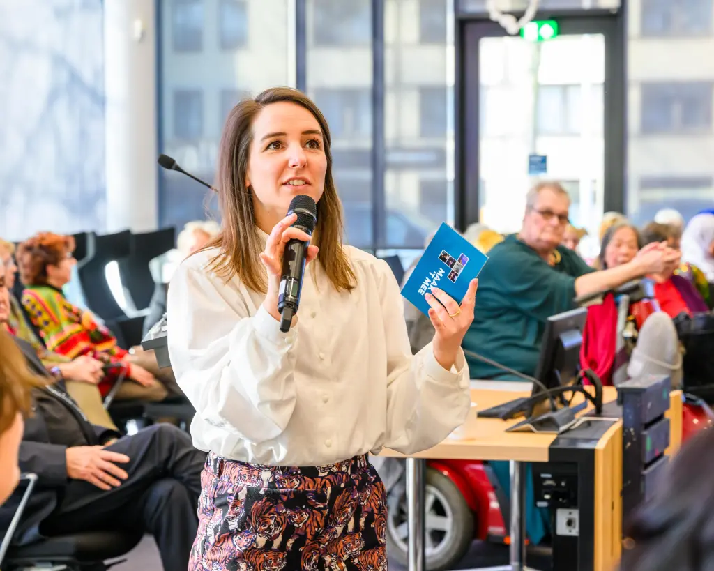 Lobke geeft een presentatie en staat temidden van haar publiek. Ze heeft een microfoon in haar hand en een stapel presentatiekaartjes. Ze draagt een witte blouse en een broek met tijgers erop.