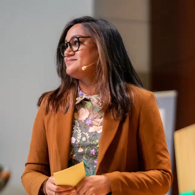 Madhu Mathoera tijdens het geven van een presentatie. Ze heeft een bruin pak aan en draagt haar haar los. In haar hand houdt ze presentatiekaartjes.