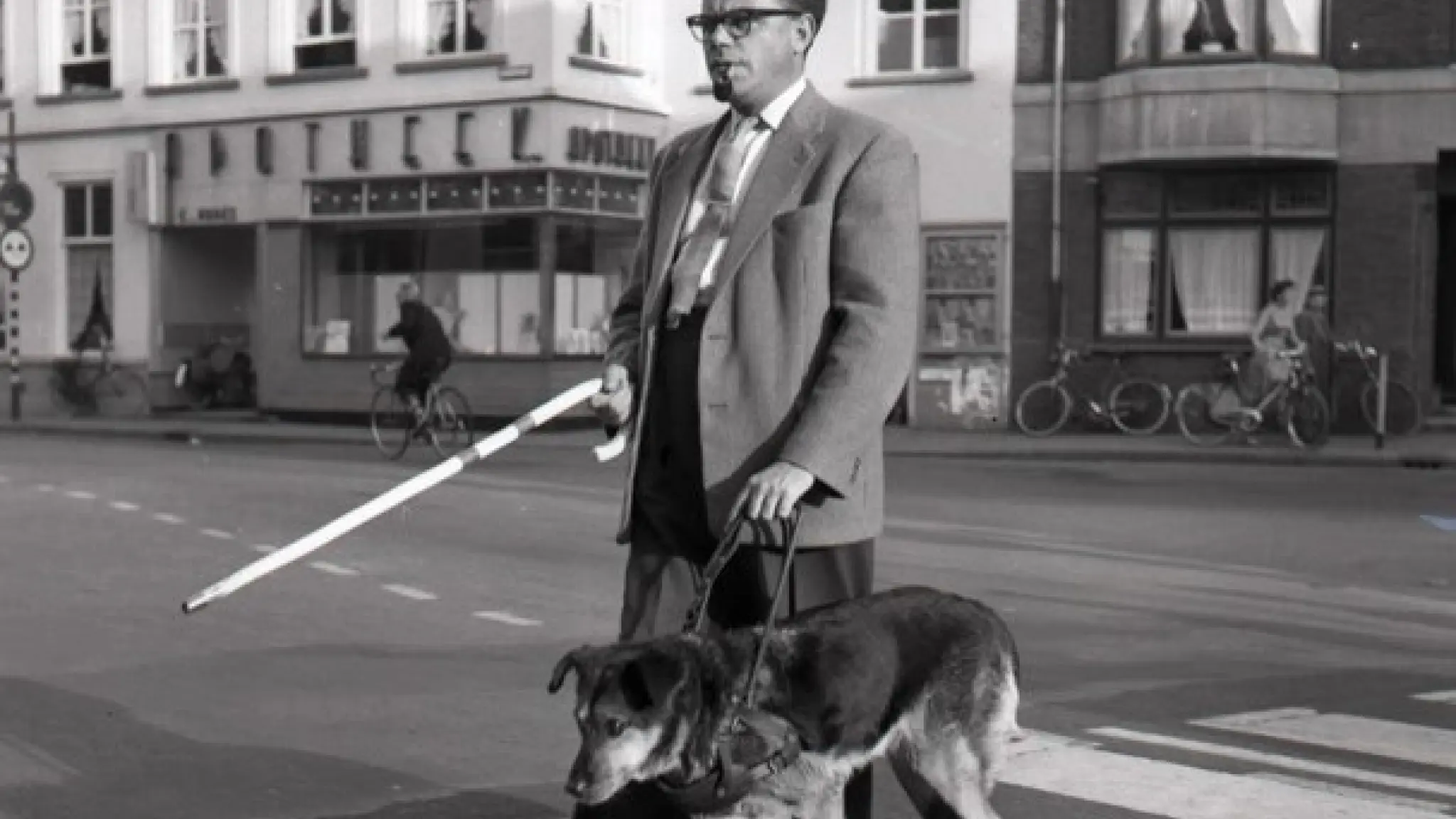 Zwart-wit foto van een man met een blindeheleidehond die een zebrapad oversteekt. Hij heeft een stok voor zich uitgestoken.