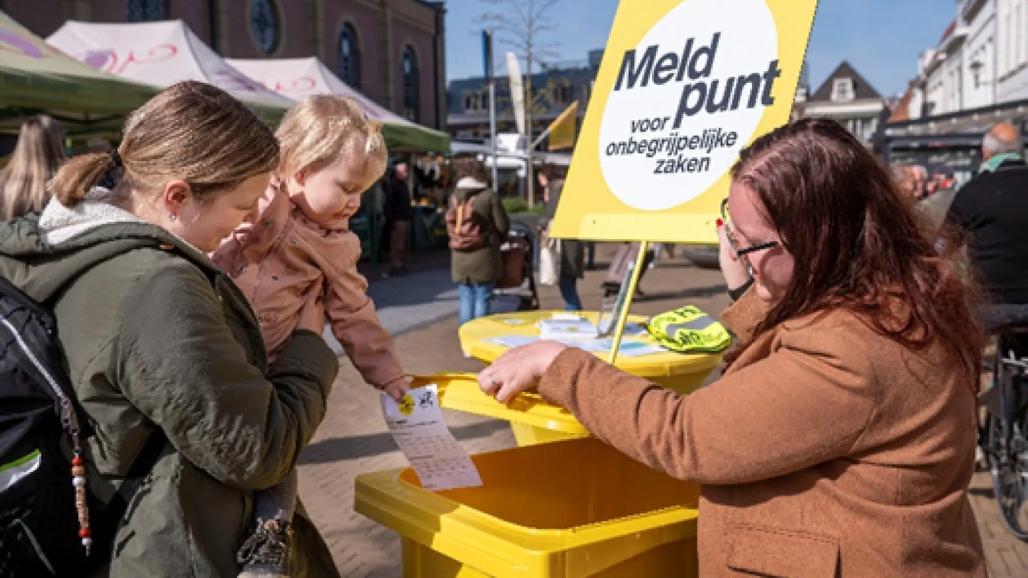 Op een marktplein staat een gele kliko met een bord Meldpunt voor onbegrijpelijke zaken. Een vrouw met een kindje op haar arm gooit een briefje in deze container.