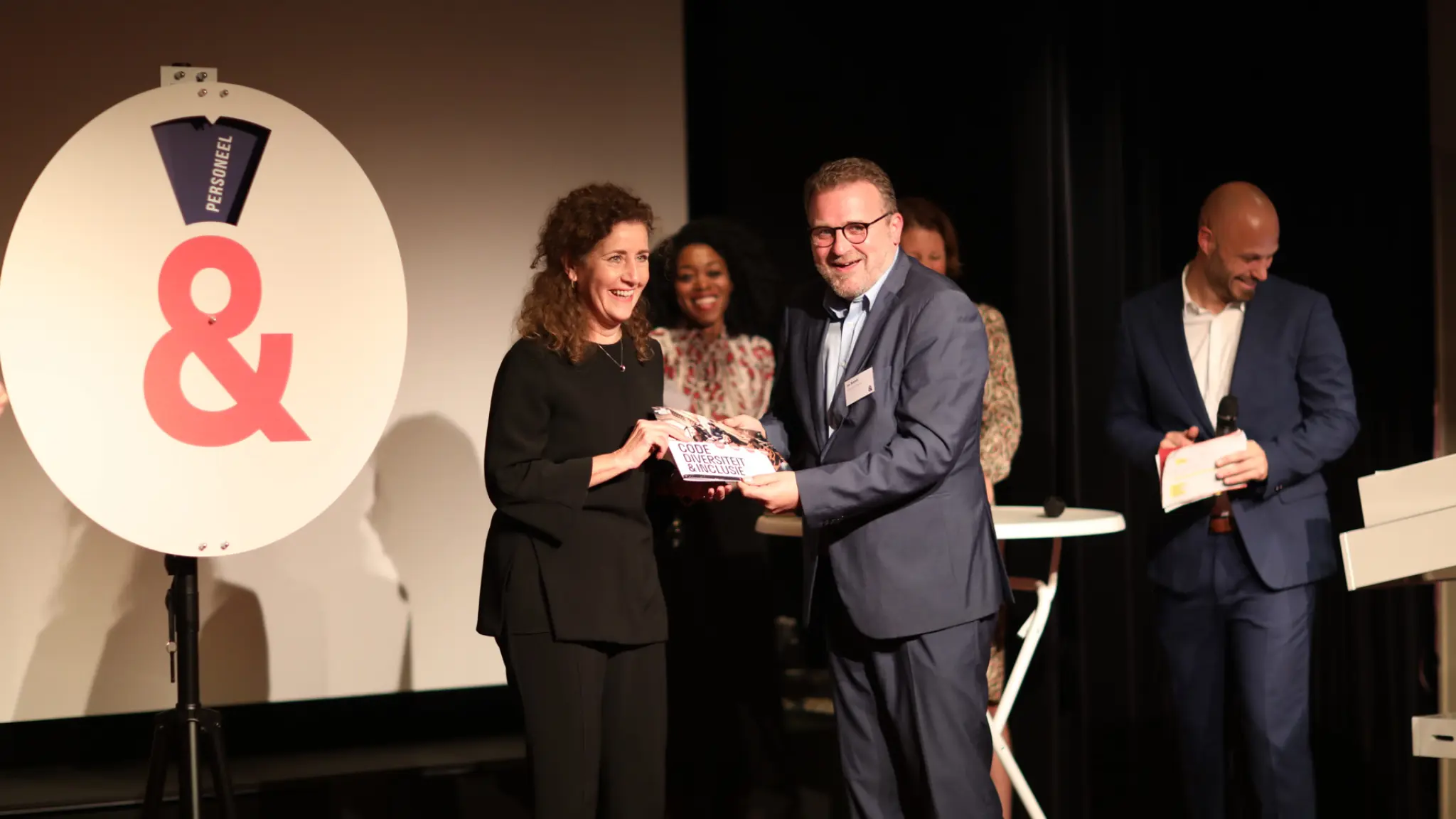 Minister Ingrid van Engelshoven op het podium voor het Pim-Pam-Pet-rad van de Code Diversiteit en Inclusie. Er staan nog vier andere mensen op het podium.