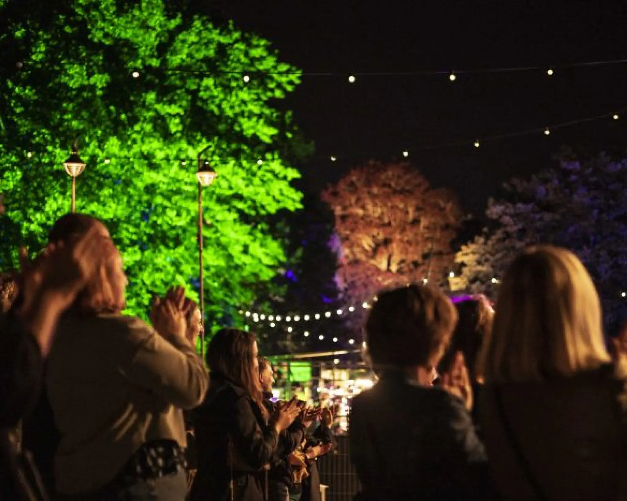 Publiek 's avonds op een sfeervol verlicht festivalterrein. Ze klappen voor een voorstelling.