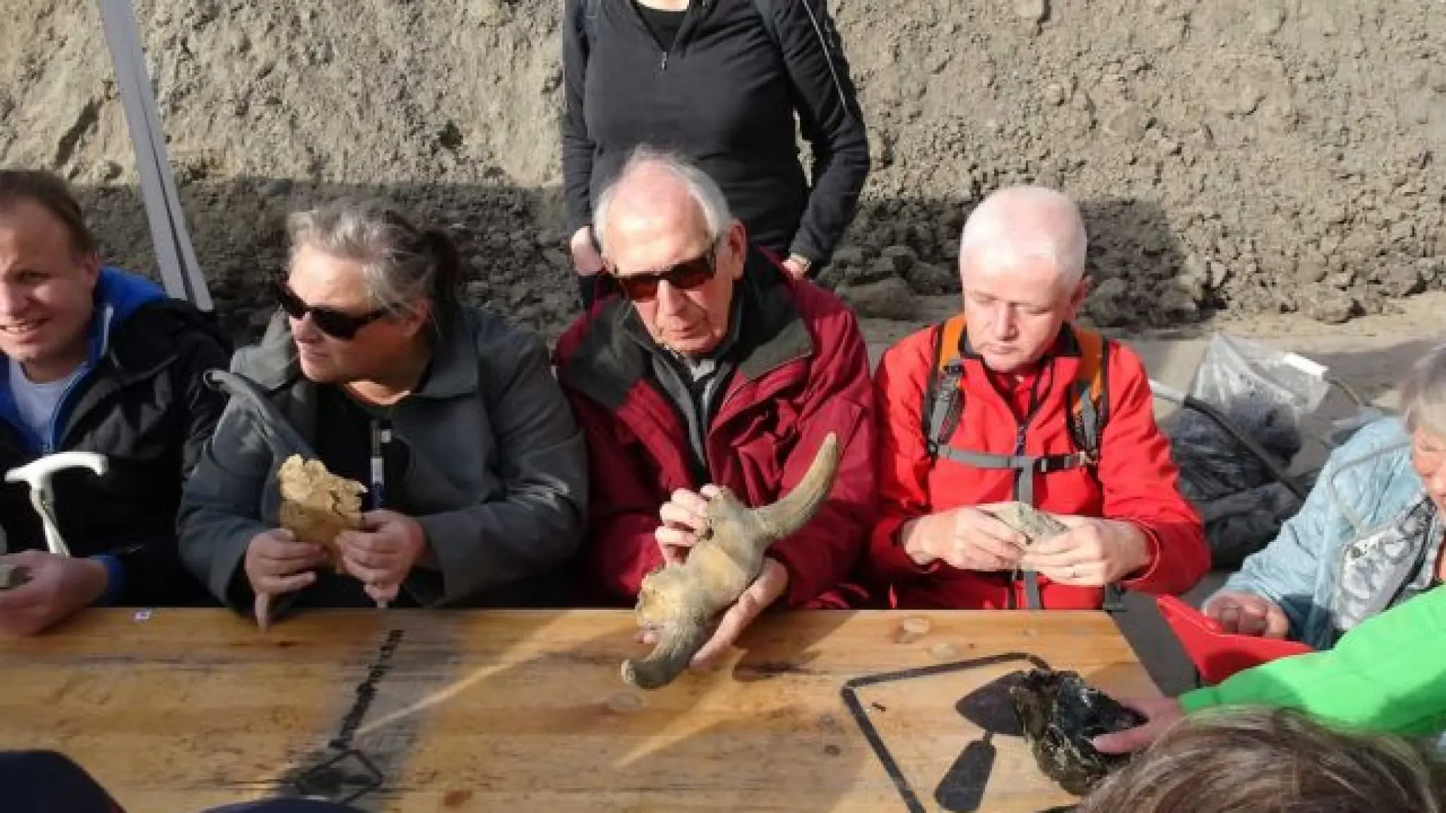 Blinde en slechtziende mensen zitten aan een tafel op een opgravingsterrein. Men hun handen voelen ze aan archeologische objecten.
