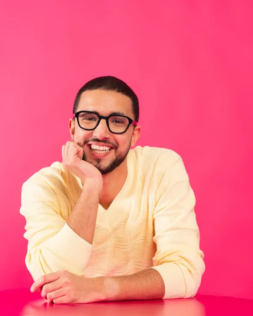 Hamza steunt met zijn elleboog op een roze tafel en leunt met zijn hoofd op zijn handpalm. Hij kijkt breed lachend naar de camera. Hij draagt een lichtgele trui en zit voor een felroze achtergrond.