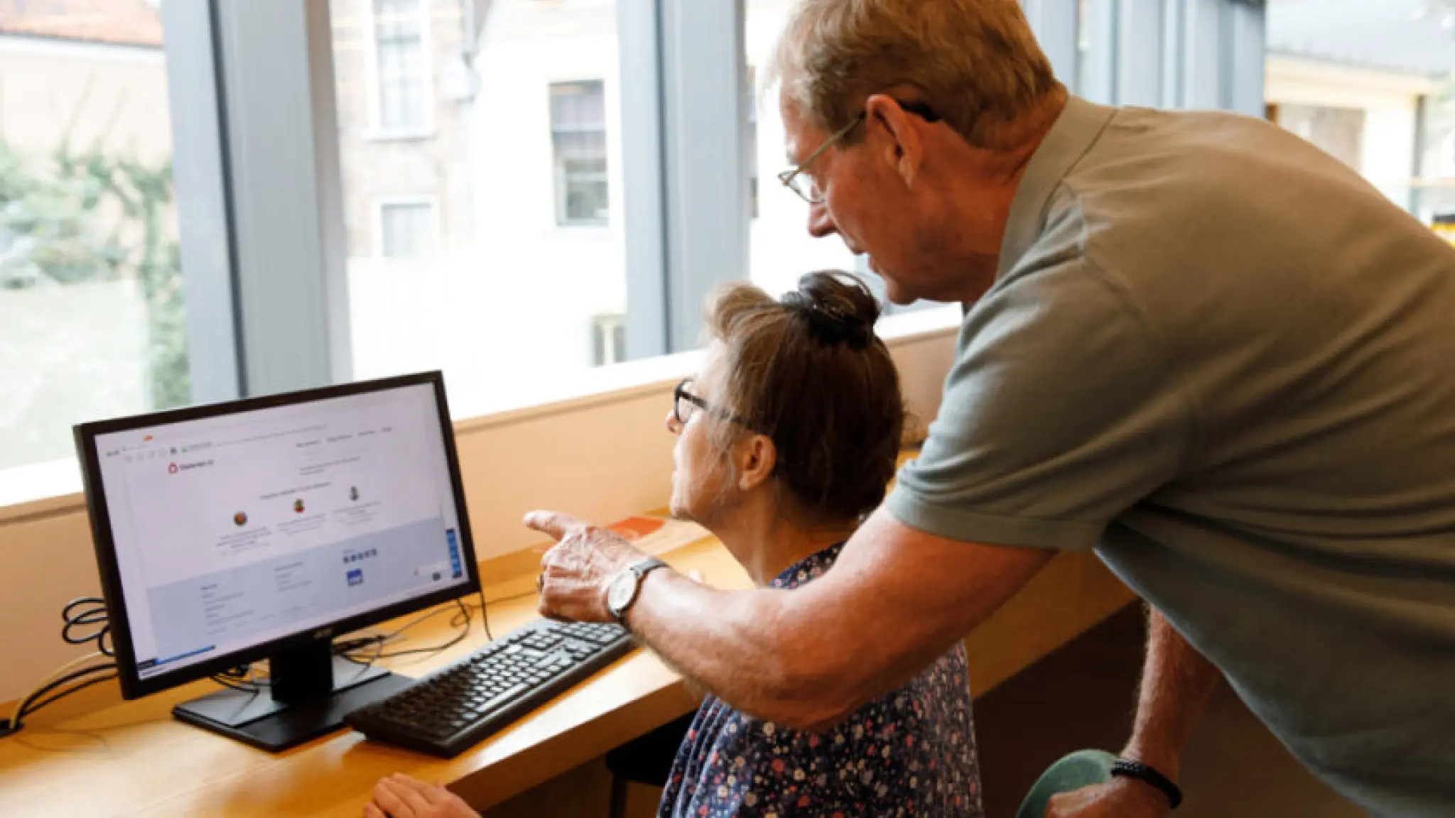 Oudere man helpt een oudere vrouw bij het gebruik van een computer in een lichte ruimte.