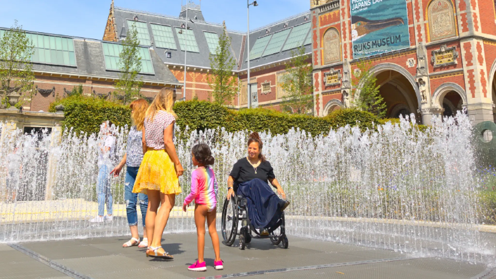 Vier bezoekers, waarvan één rolstoelgebruiker, staan tussen opspuitende waterfonteinen op het Museumplein in Amsterdam. Op de achtergrond is de ingang van het Rijksmuseum zichtbaar.