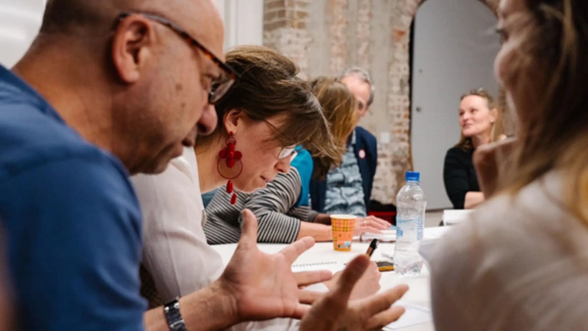 Deelnmers aan de training zitten met z'n allen rond een tafel en praten in twee- en drietallen met elkaar. Op tafel liggen papiere, , boekjes, pennen, een koffiebeker en een flesje water. Op de achtergrond is een stuk van een bakstenen muur te zien.
