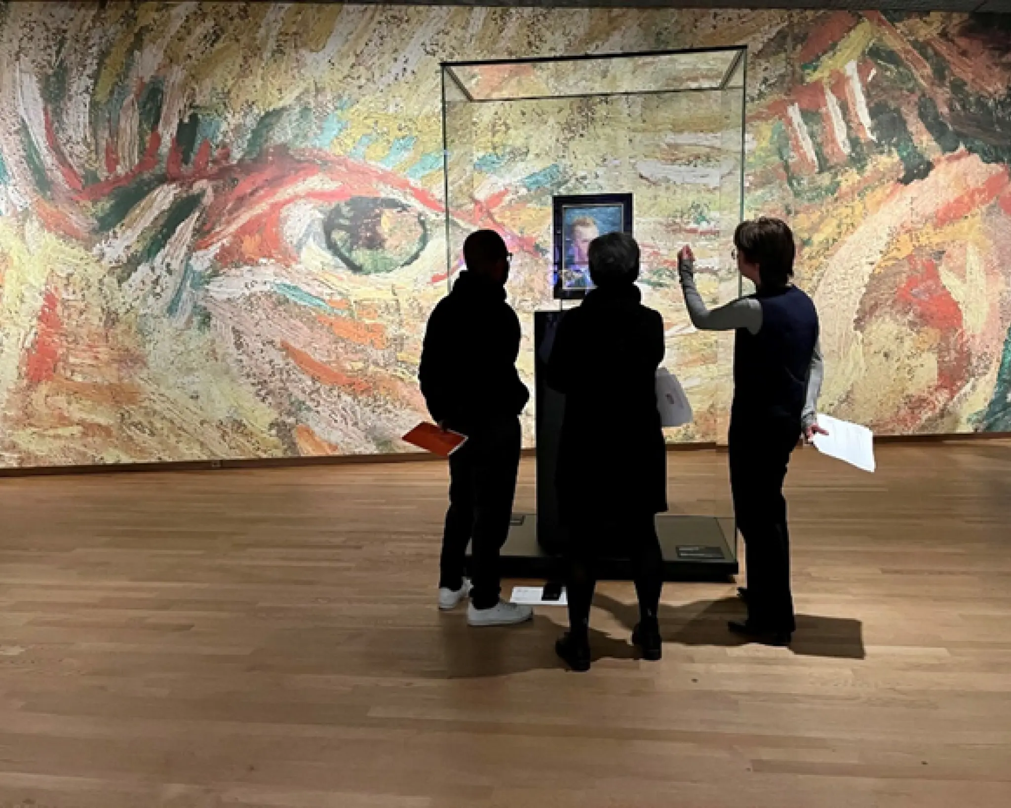 Drie mensen staan voor een vitrine met daarin een schilderij van Van Gogh. Op de achtergrond is een wand te zien met daarop een deel van een van de schilderijen van Van Gogh. Je ziet een oog en een oor.