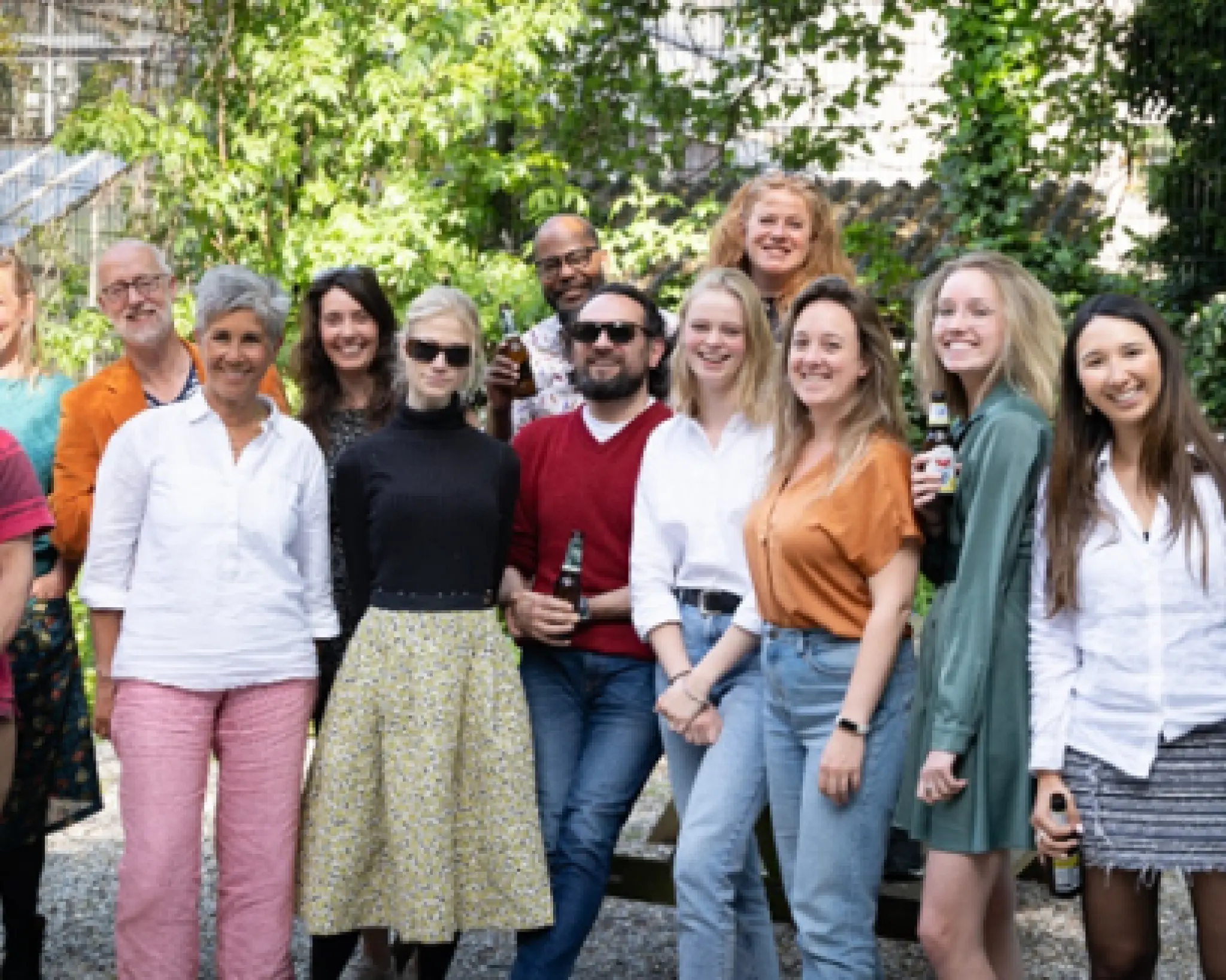 Een groep mensen poseert lachend voor een groepsfoto. Ze staan onder een boom. Sommigen houden een bierflesje in hun hand.