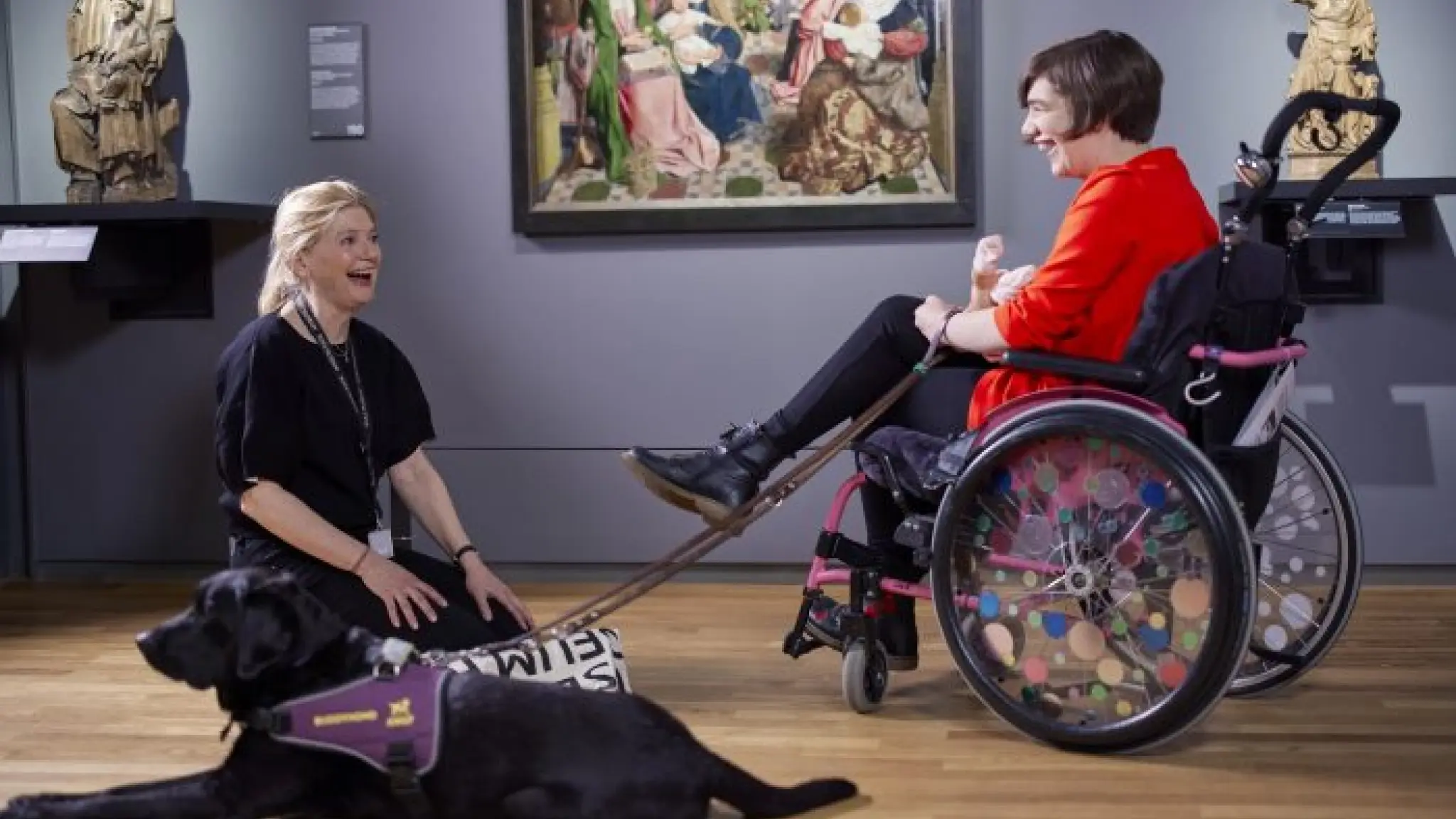 Een jonge vrouw zit in een rolstoel in een galerij van het Rijksmuseum. Ze heeft een hulphond bij zich die naast haar op de grond ligt. Een rondleidster zit op haar knieën op de grond naast de hond. Beide vrouwen lachen.