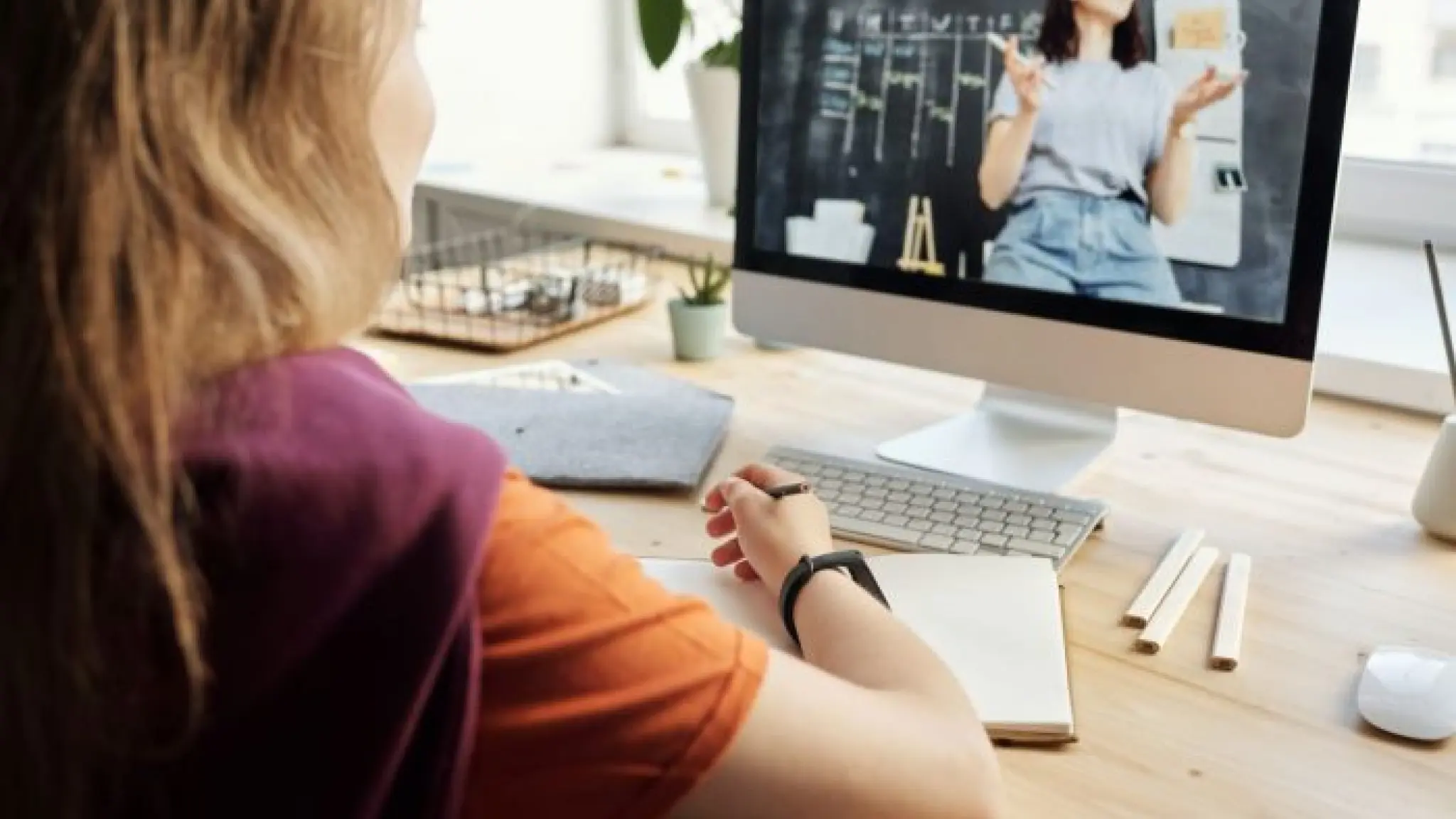 Een vrouw kijkt naar een online uitleg op haar pc. Ze heeft een opschrijfboekje open voor haar liggen en een potlood in haar hand.