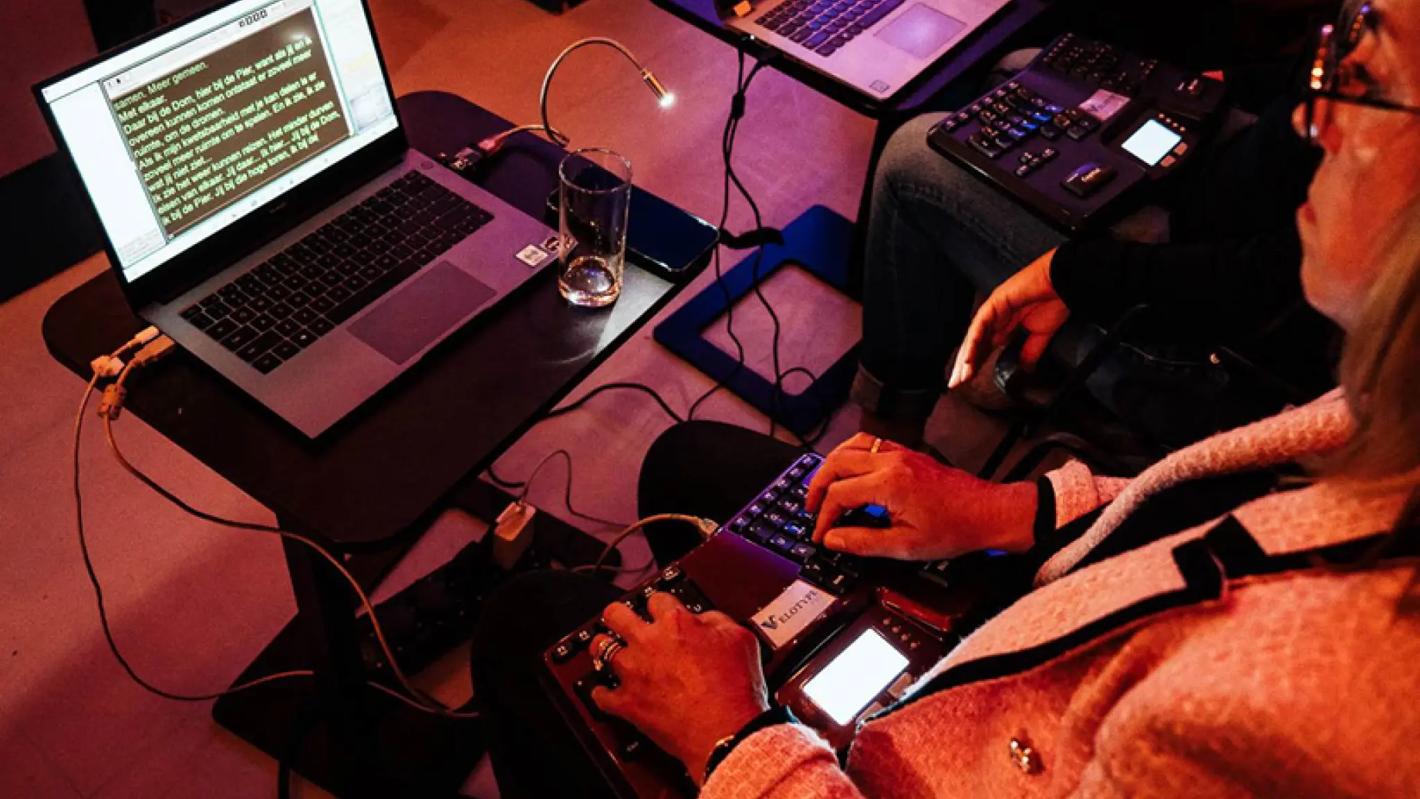 Twee mensen zitten achter laptops en stenografiemachines, waarbij op het laptopscherm realtime ondertiteling in gele tekst op een zwarte achtergrond te zien is. Een glas water en een kleine leeslamp staan op het tafeltje naast één van de laptops.