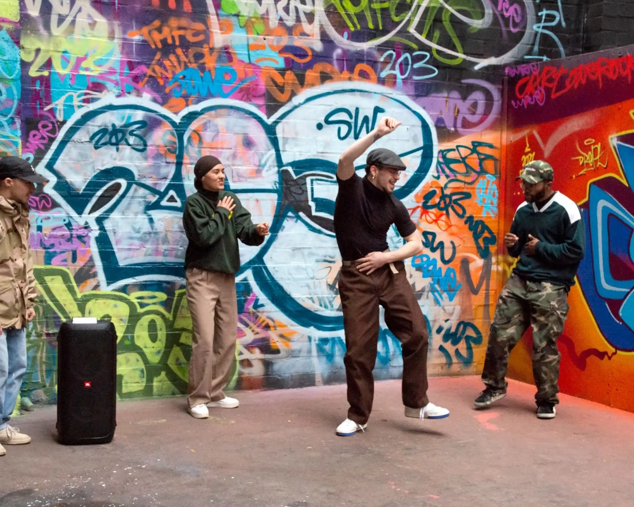 Vier mensen dansen op een met graffiti bespoten binnenplaats. Op de grond staat een verrijdbare muziekbox.