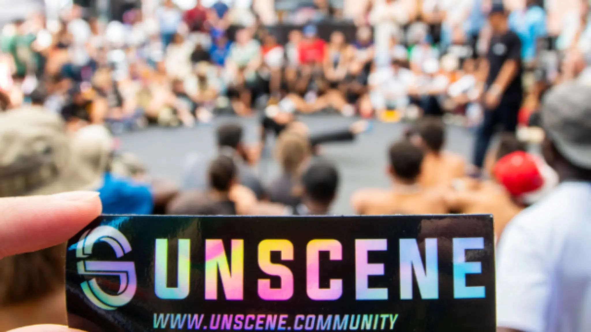 Een hand houdt een sticker vast met daarop in kleurige letters UNSCENE en www.unscene.community. Op de achtergrond is een cirkel van mensen te zien met in het midden twee mensen bezig met een break dance battle.