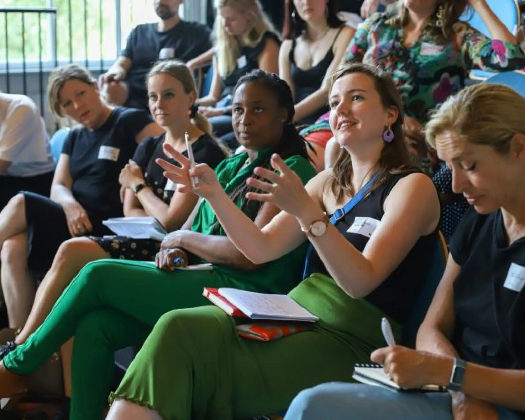 Bezoekers van de bijeenkomst zitten op een tribune. Ze dragen zomerse kleding. Een vrouw op de eerste rij is in gesprek met iemand buiten beeld en heeft haar handen opgeheven alsof ze denkbeeldig iets voor zich vasthoudt.