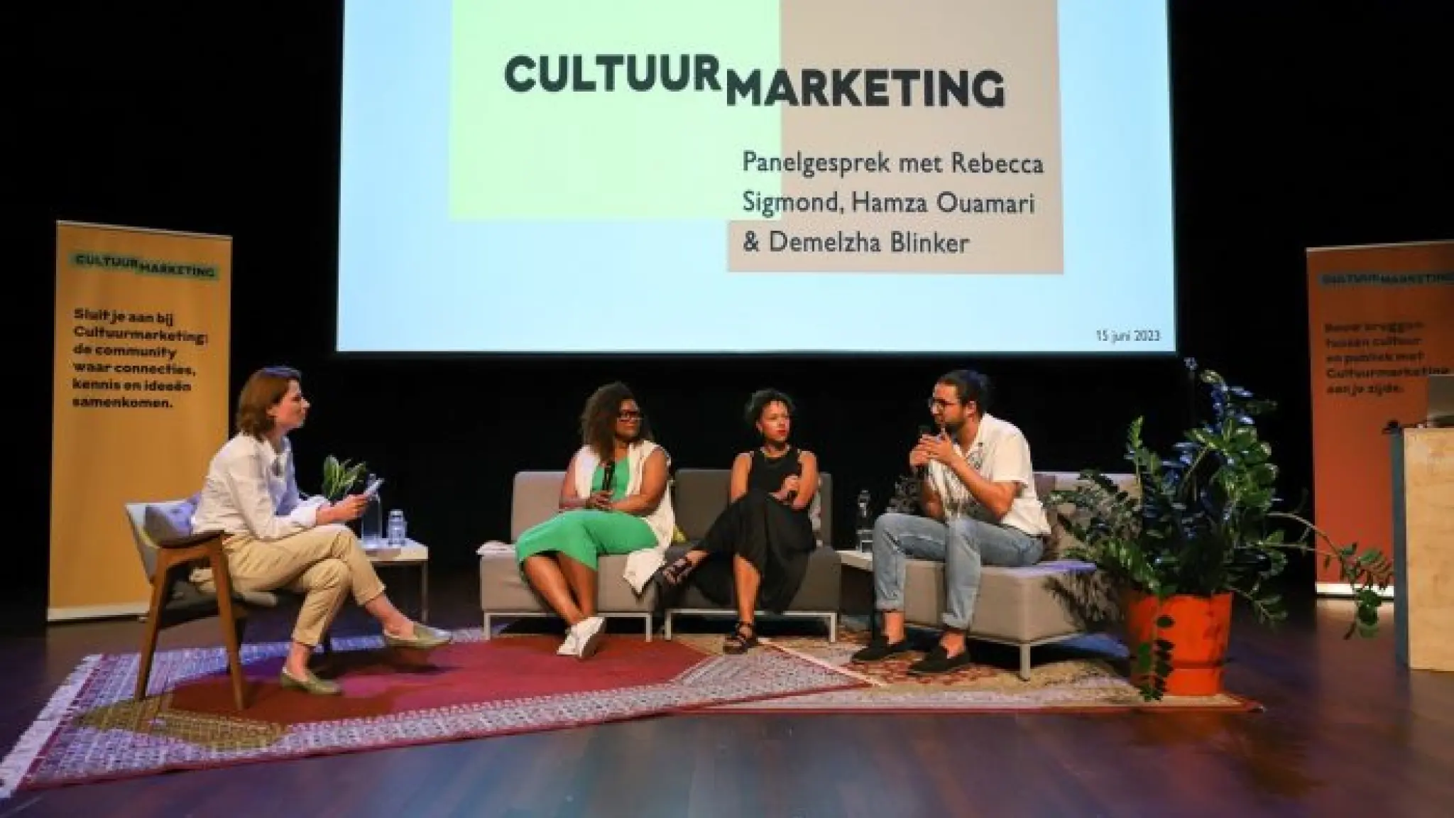 Vier mensen zitten op een podium en zijn met elkaar in gesprek. Het podium is aangekleed als huiskamer, met comfortabele banken en stoelen, kleden en kamerplanten. Links en rechts staan banieren van Cultuurmarketing. Achter hen hangt een groot scherm met daarop Cultuurmarketing - panelgesprek met Rebecca Sigmond, Hamza Ouamari & Demelzha Blinker.