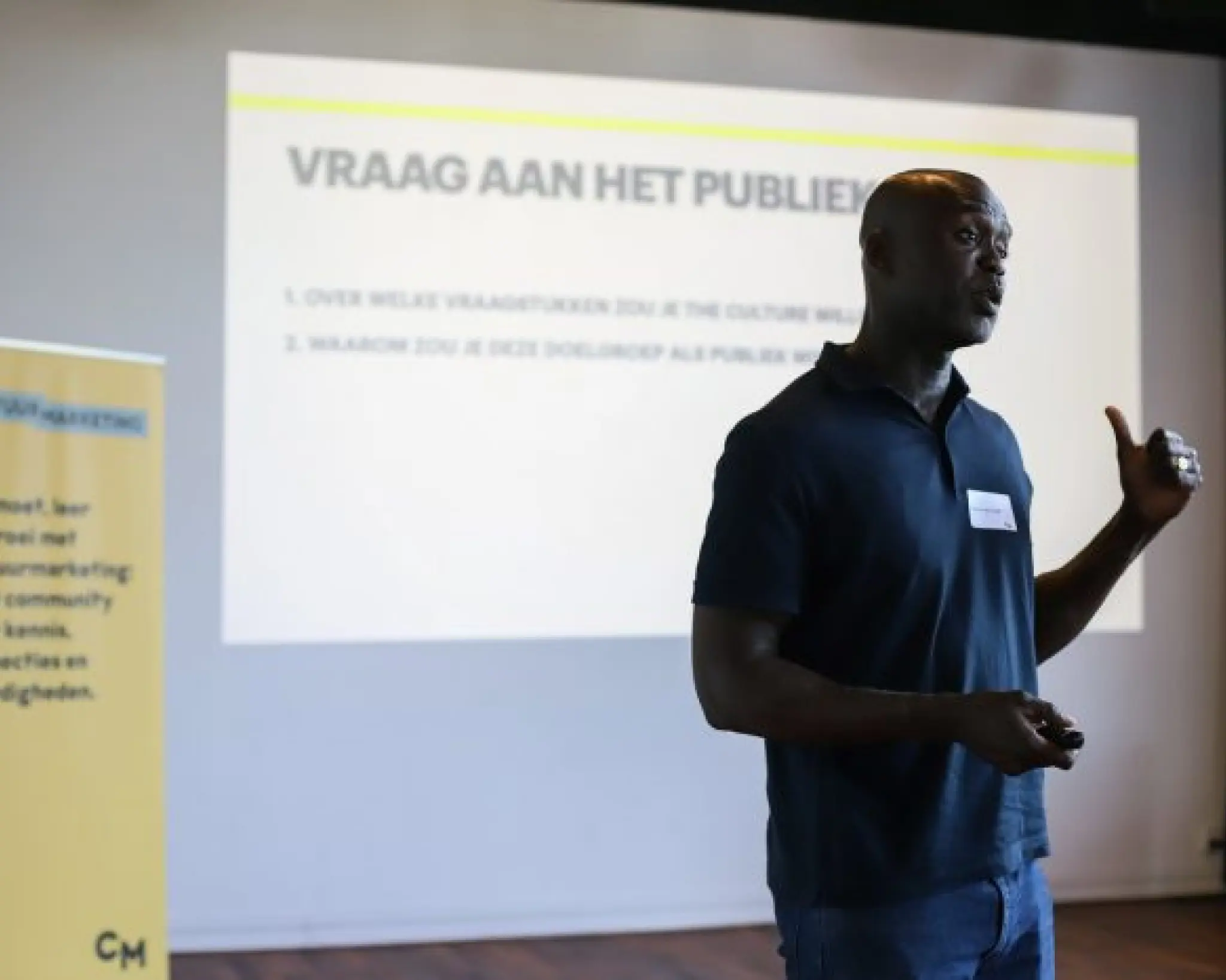 Een donkere man staat voor een presentatiescherm met daarop de tekst 'Vraag aan het publiek'. Links van hem staat een banier van Cultuurmarketing.
