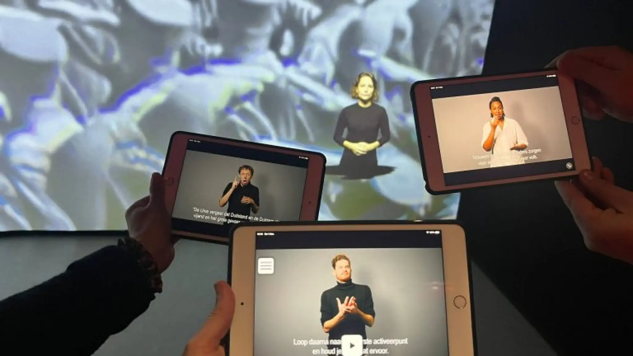 Drie handen houden een tablet vast waarop een video met een tolk Nederlandse Gebarentaal is te zien. Op de achtergrond is een groot videoscherm van het museum te zien met daarop ook een tolk NGT.