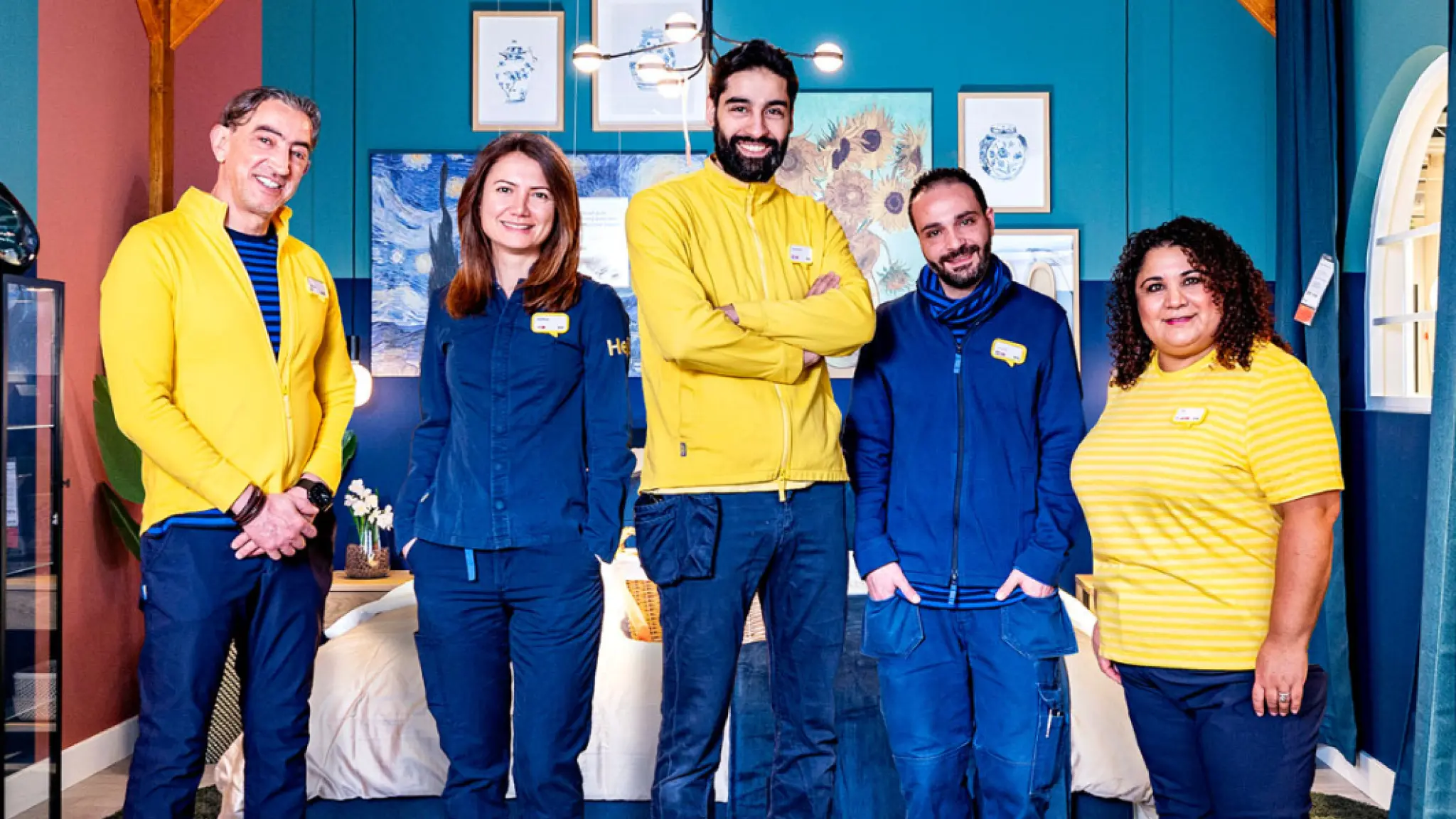 Vijf IKEA-medewerkers staan lachend naast elkaar in een ingerichte slaapkamer. Ze dragen blauwe of gele bedrijfskleding en poseren voor een kleurrijke muur met schilderijen.