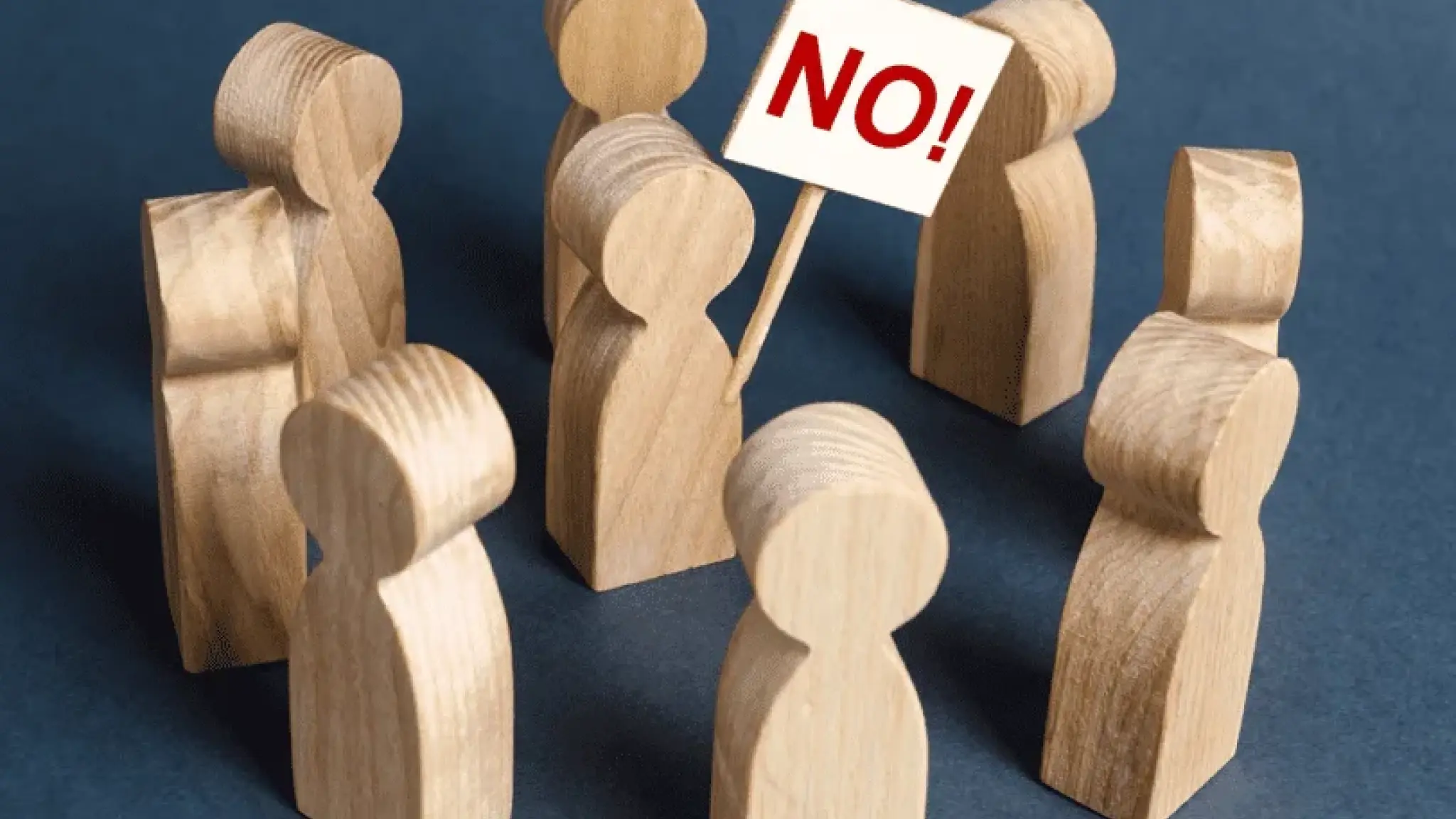 Een groep houten poppetjes staat in een cirkel, waarbij één poppetje een bord vasthoudt met de tekst "NO!" erop.