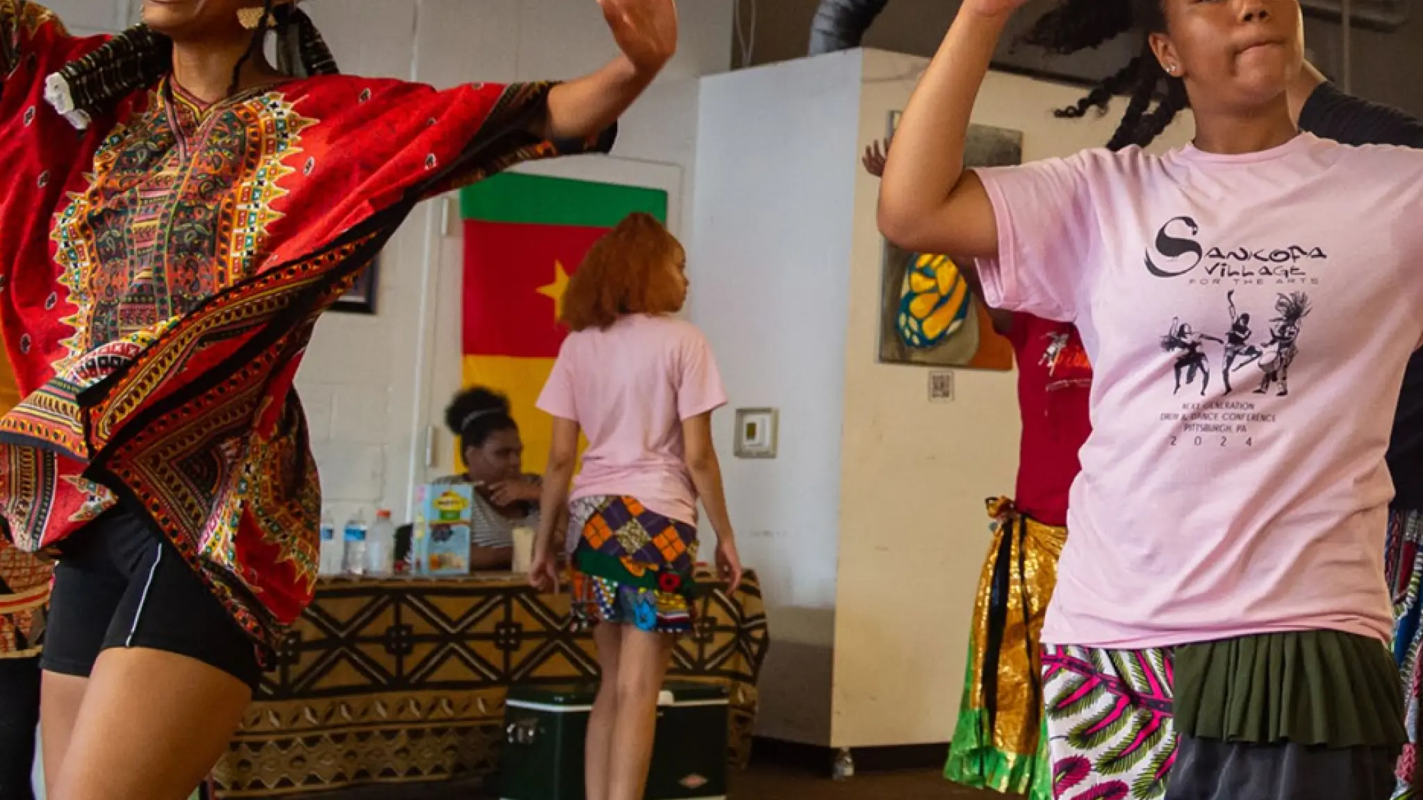 Twee jonge vrouwen dansen vrolijk in kleurrijke kleding in een binnenruimte, met op de achtergrond andere mensen en een vlag aan de muur.